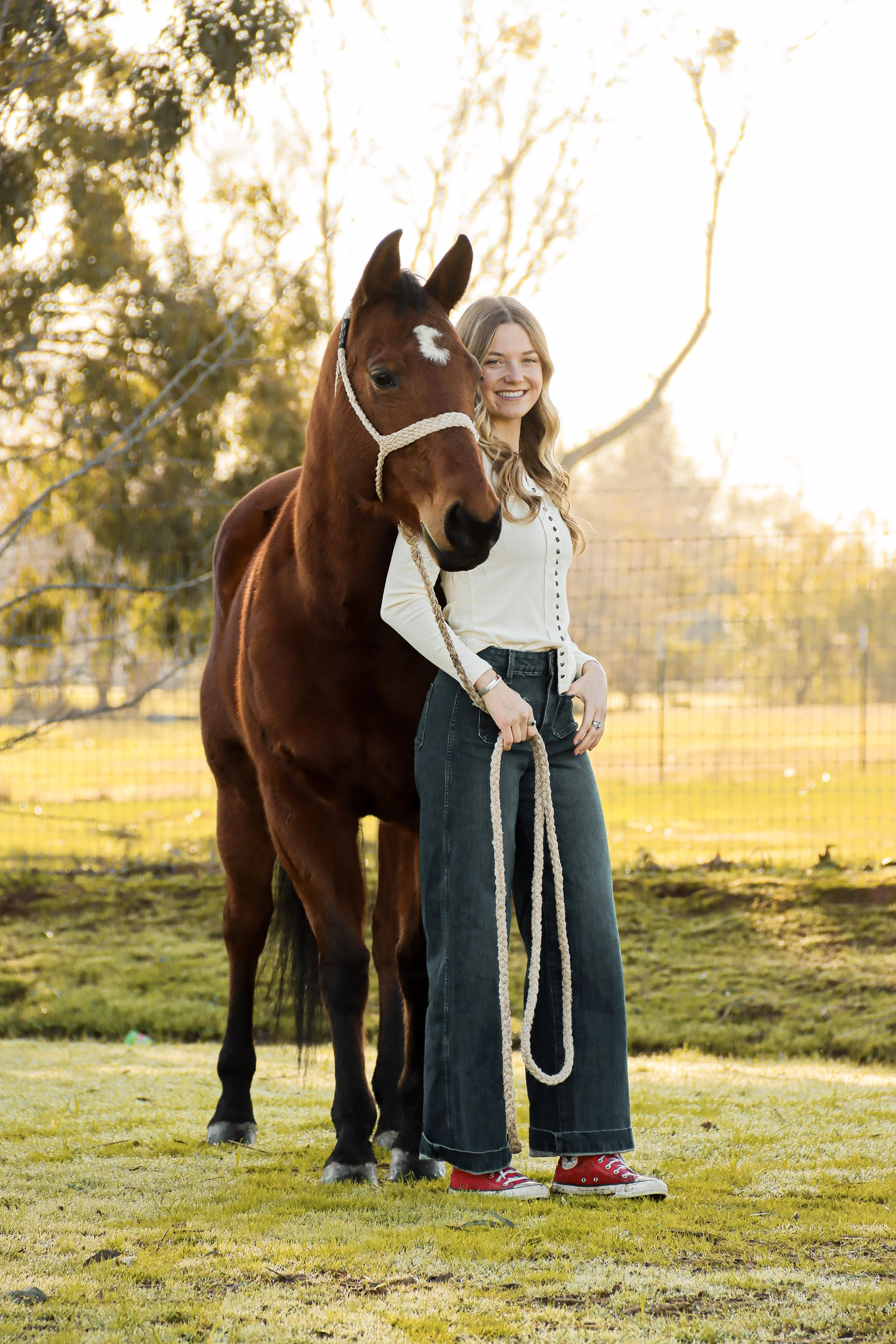 A young woman with long, wavy blonde hair stands outdoors in a grassy area with trees in the background during sunset, smiling while holding a rope attached to a brown horse with a white star-shaped mark on its forehead. The woman is wearing a white 