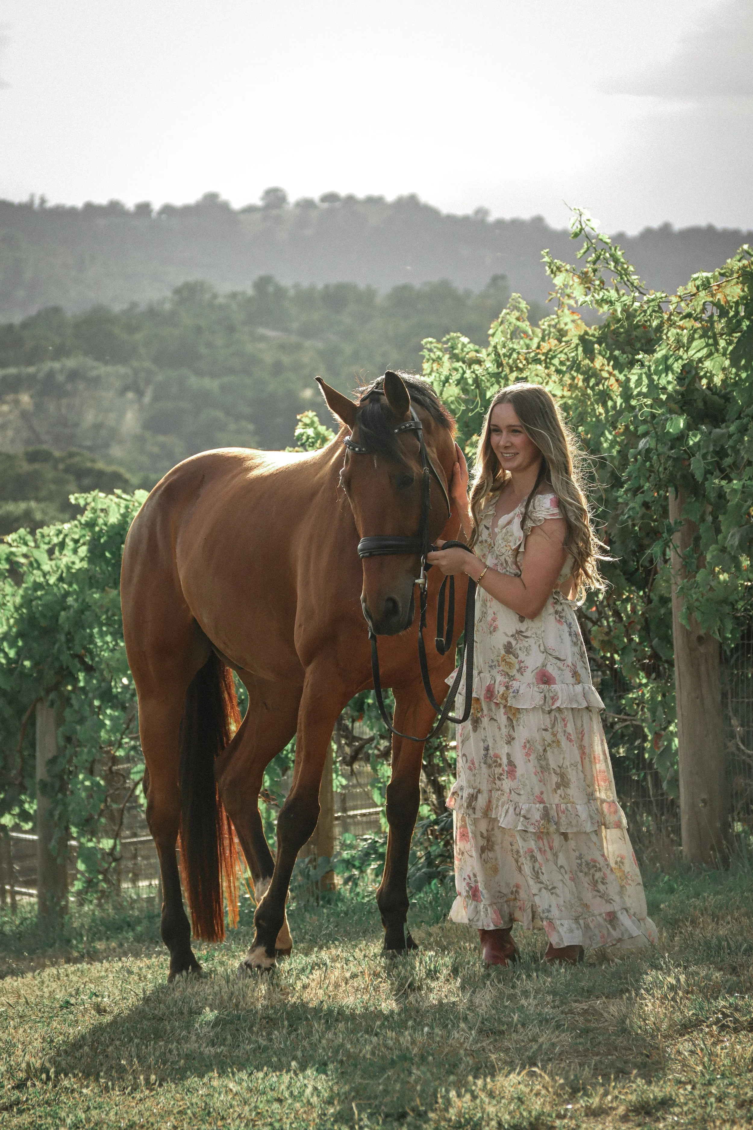 A young woman with long, wavy hair in a floral dress gently touches a brown horse's face amid lush greenery, with a mountainous landscape in the background.