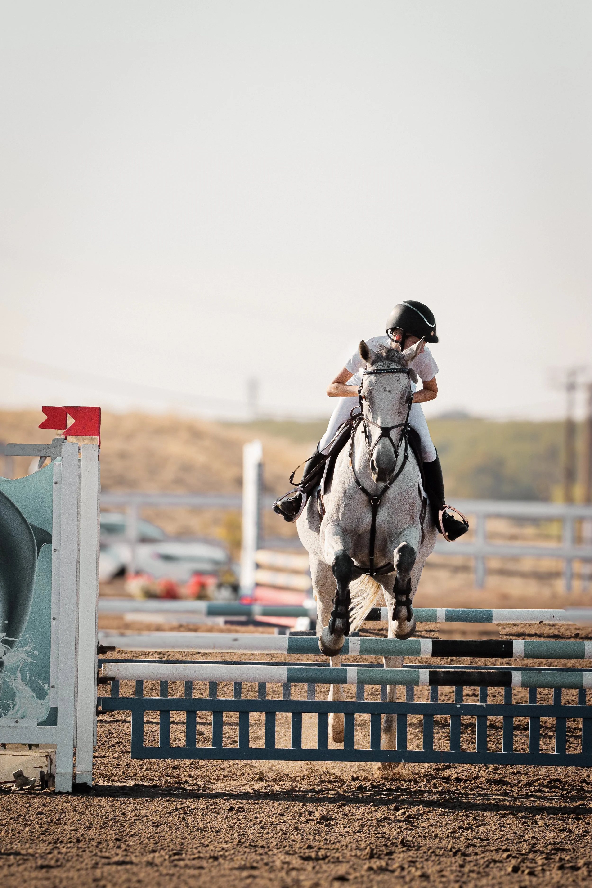 A person wearing a helmet and riding a grey horse over a show jumping obstacle in an outdoor equestrian arena.