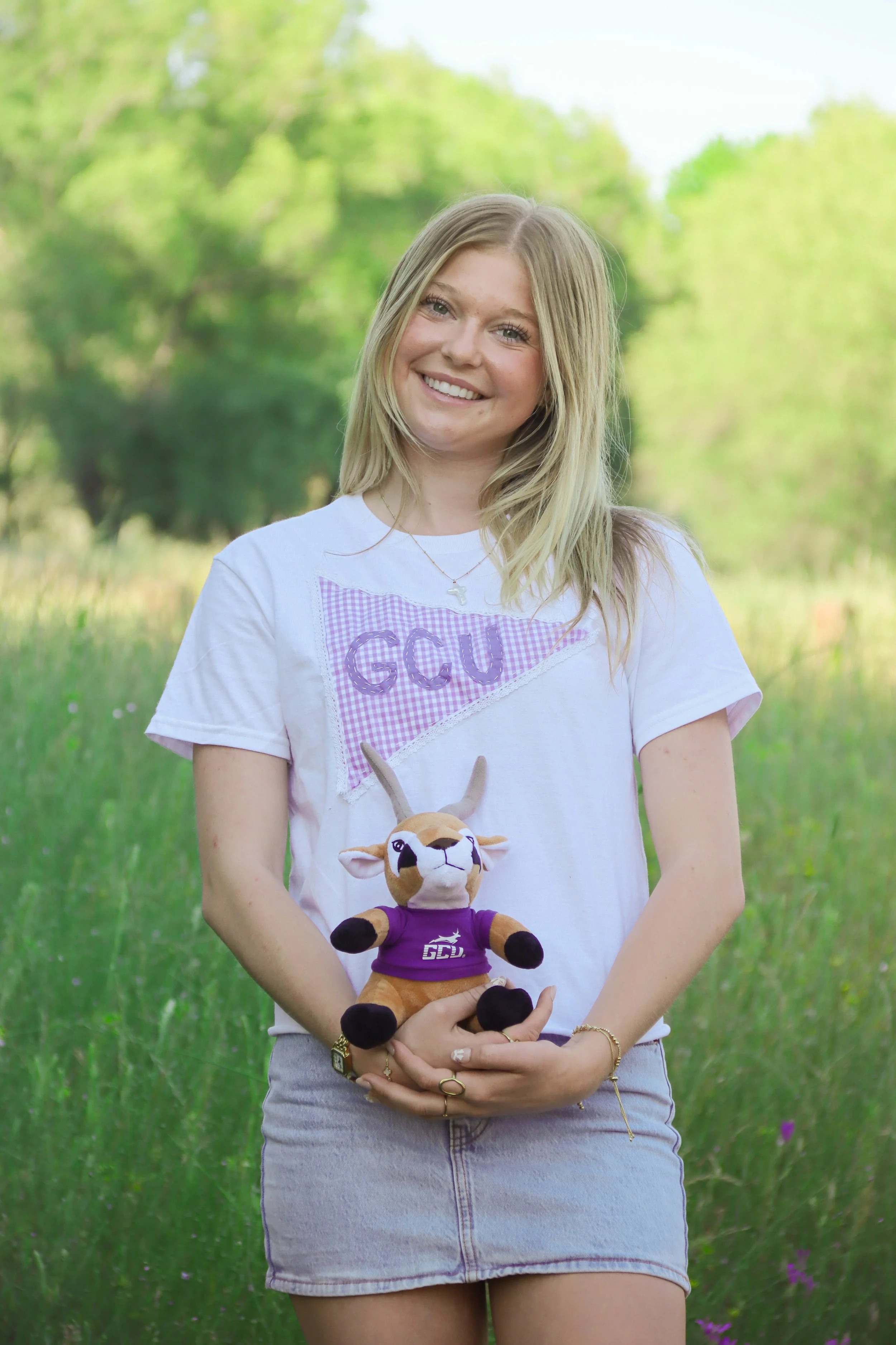 Smiling young woman in a white GC shirt holding a plush animal toy outdoors in a grassy, green park with trees.