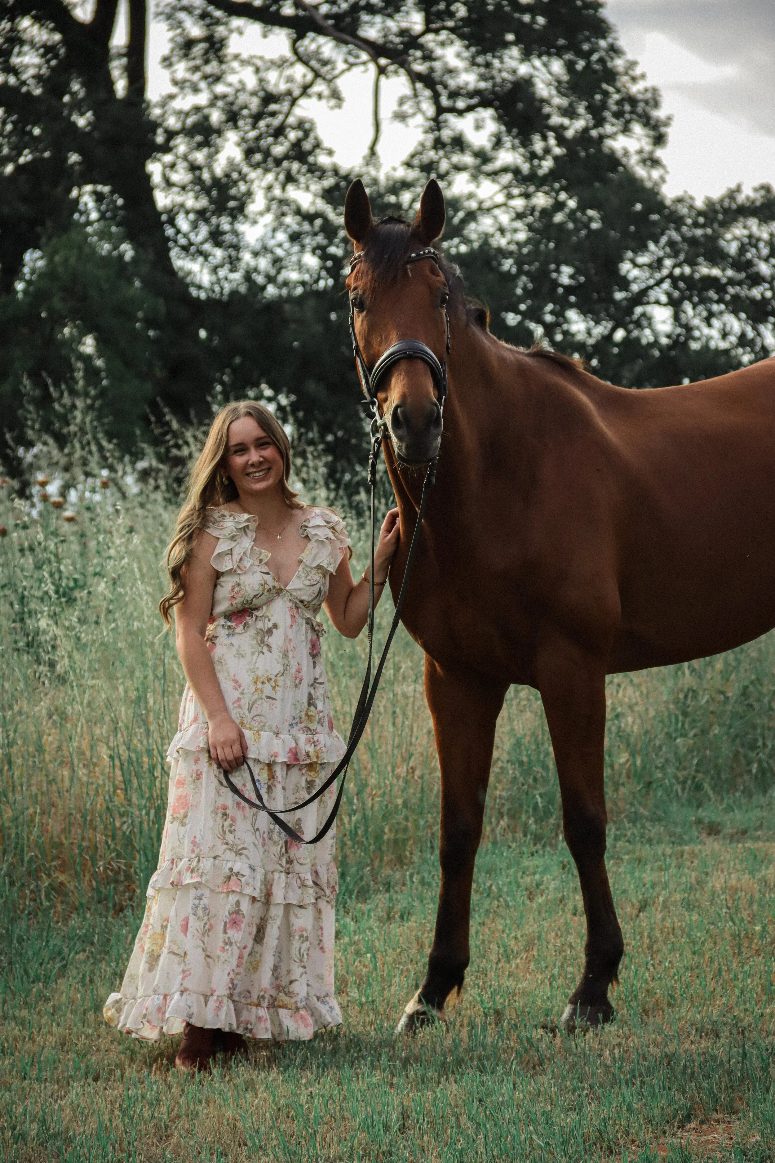 A young woman with long, wavy blonde hair, wearing a light-colored, floral, tiered dress, stands on grass holding the reins of a brown horse with a glossy coat, tall stature, and a black bridle, in a field with tall grass and wildflowers, under a lar