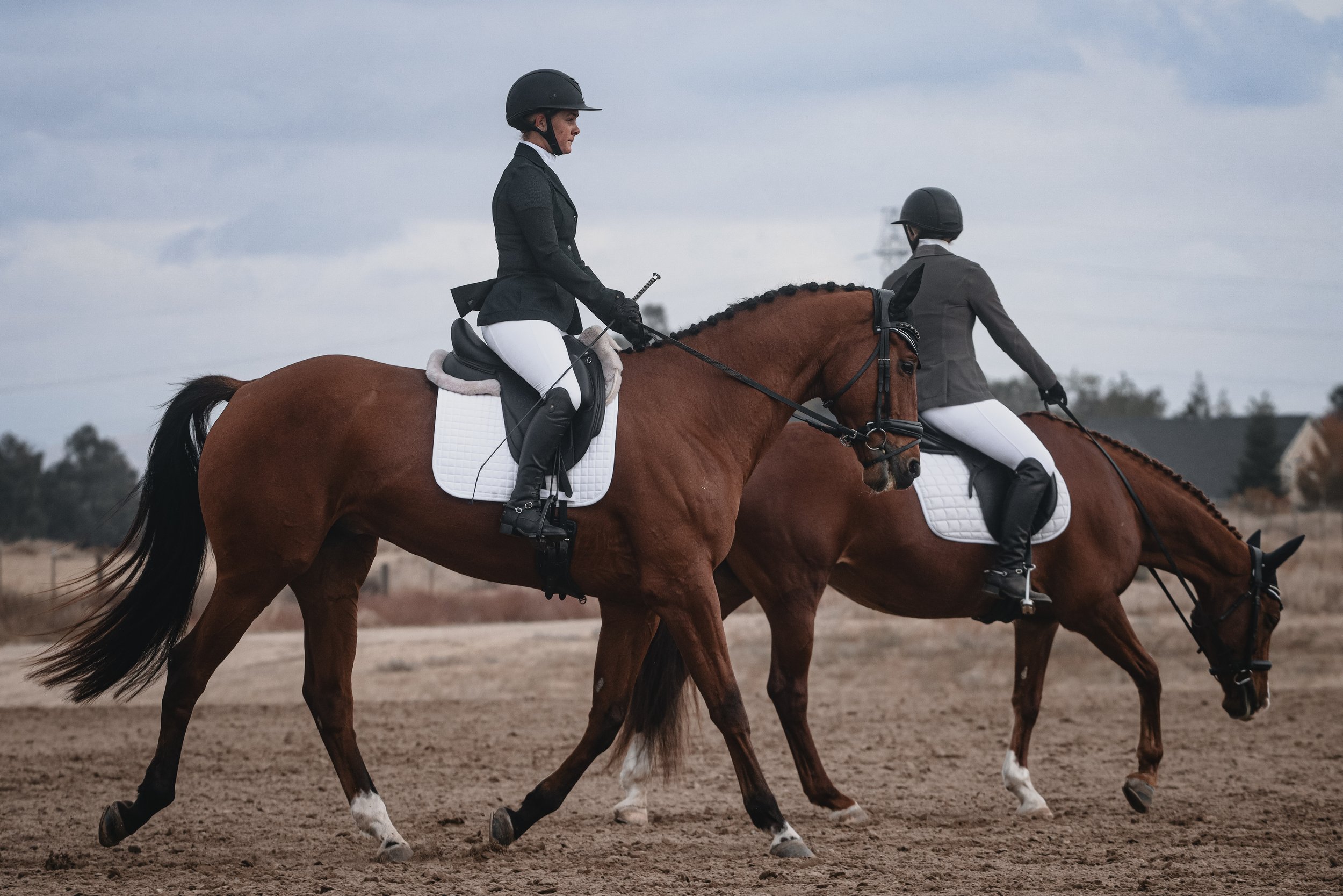 Two women riding horses on a dirt riding arena outdoors, wearing equestrian attire and helmets, with cloudy sky overhead.