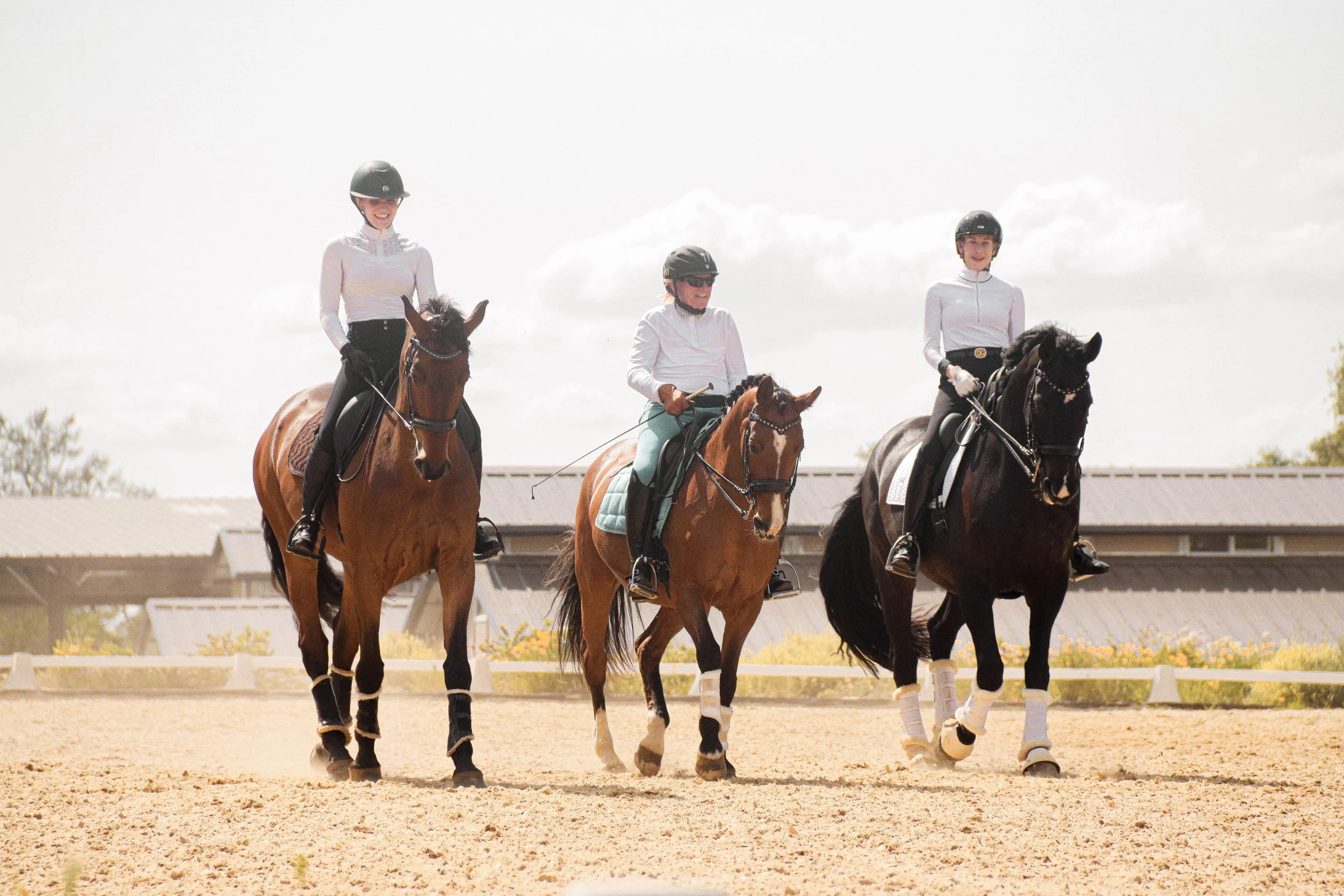 Three women riding horses in an outdoor riding arena on a sunny day, wearing helmets, riding gloves, and white riding shirts.