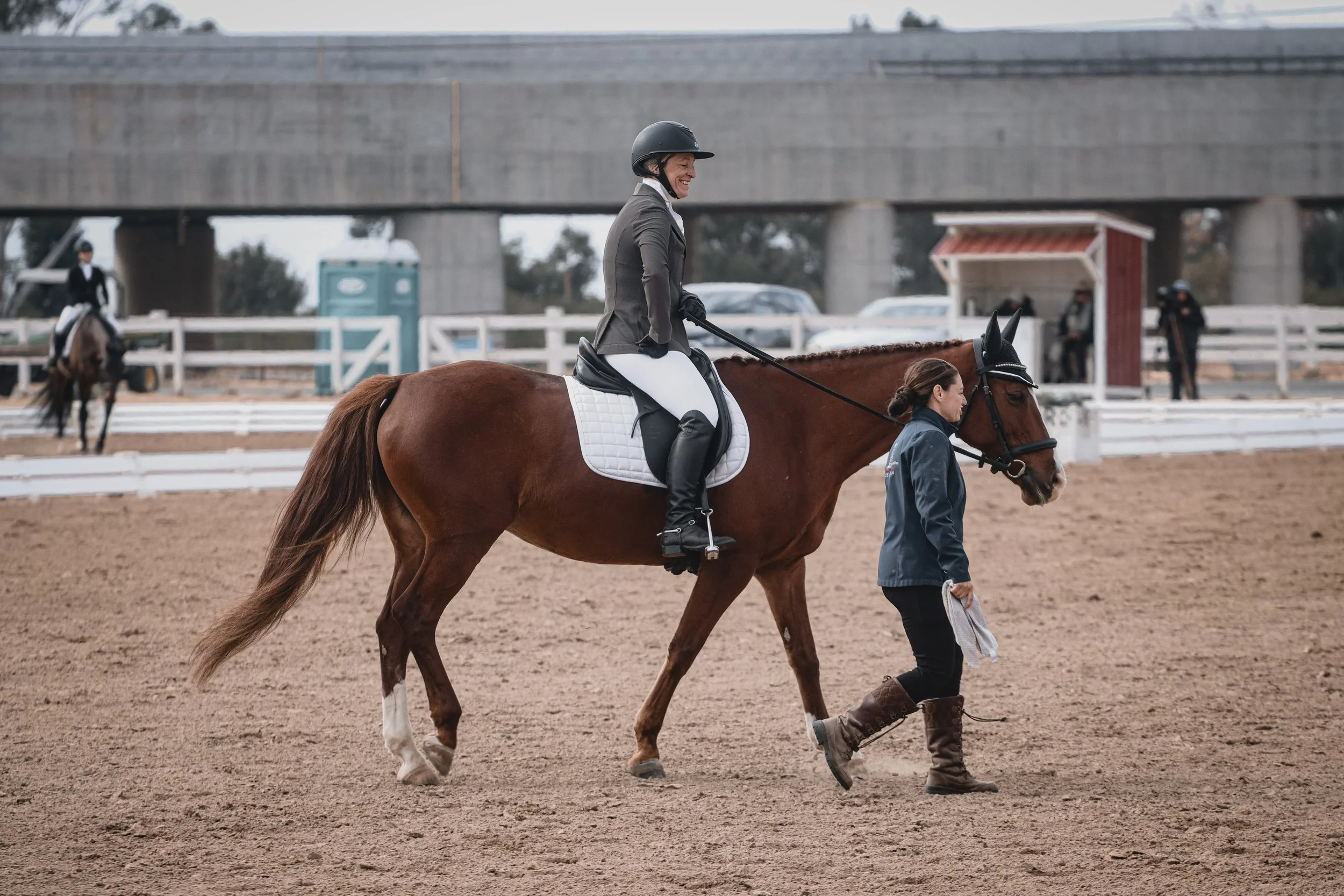 A woman wearing riding gear and a helmet is riding a brown horse in an equestrian arena. A staff member in a jacket is walking alongside the horse, guiding it. There are other riders and people in the background, with a large concrete structure and p