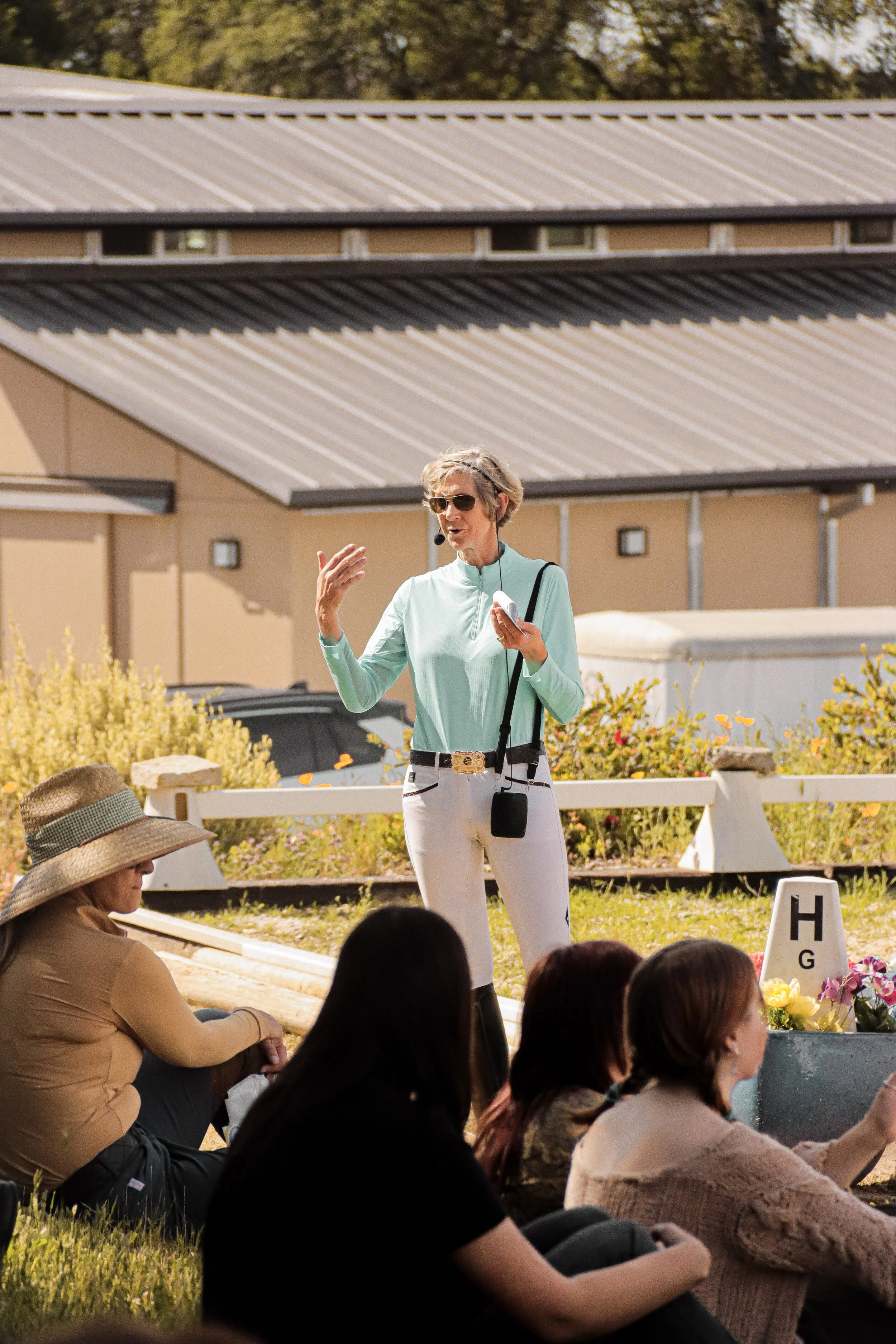 A woman wearing sunglasses, a mint green jacket, and white pants is speaking to a group of people while holding a microphone and a phone outdoors. Several women are seated on the grass, listening to her, with some wearing hats and glasses. There is a