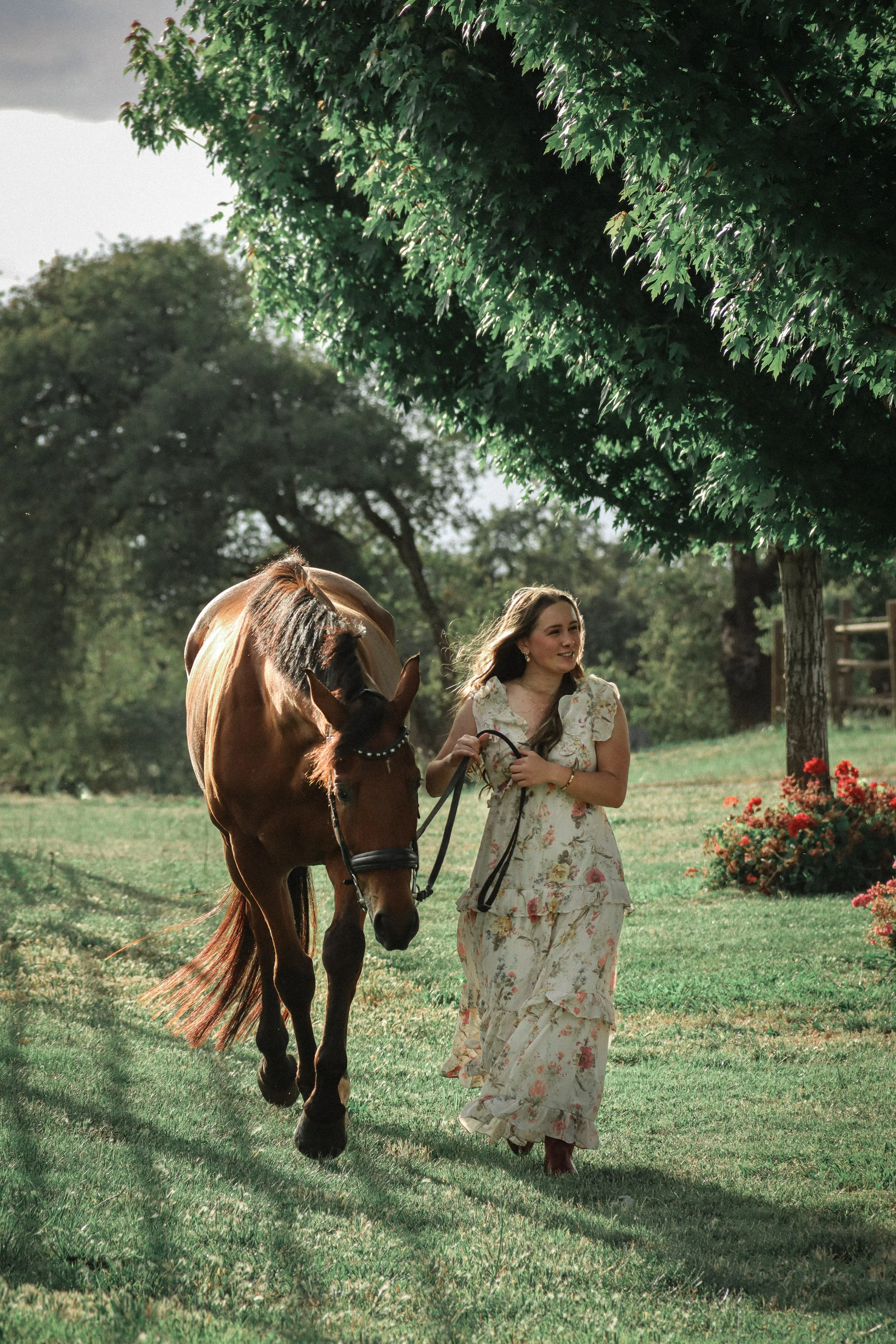 A young woman in a floral dress walking a brown horse on a leash in a park with green grass and trees.