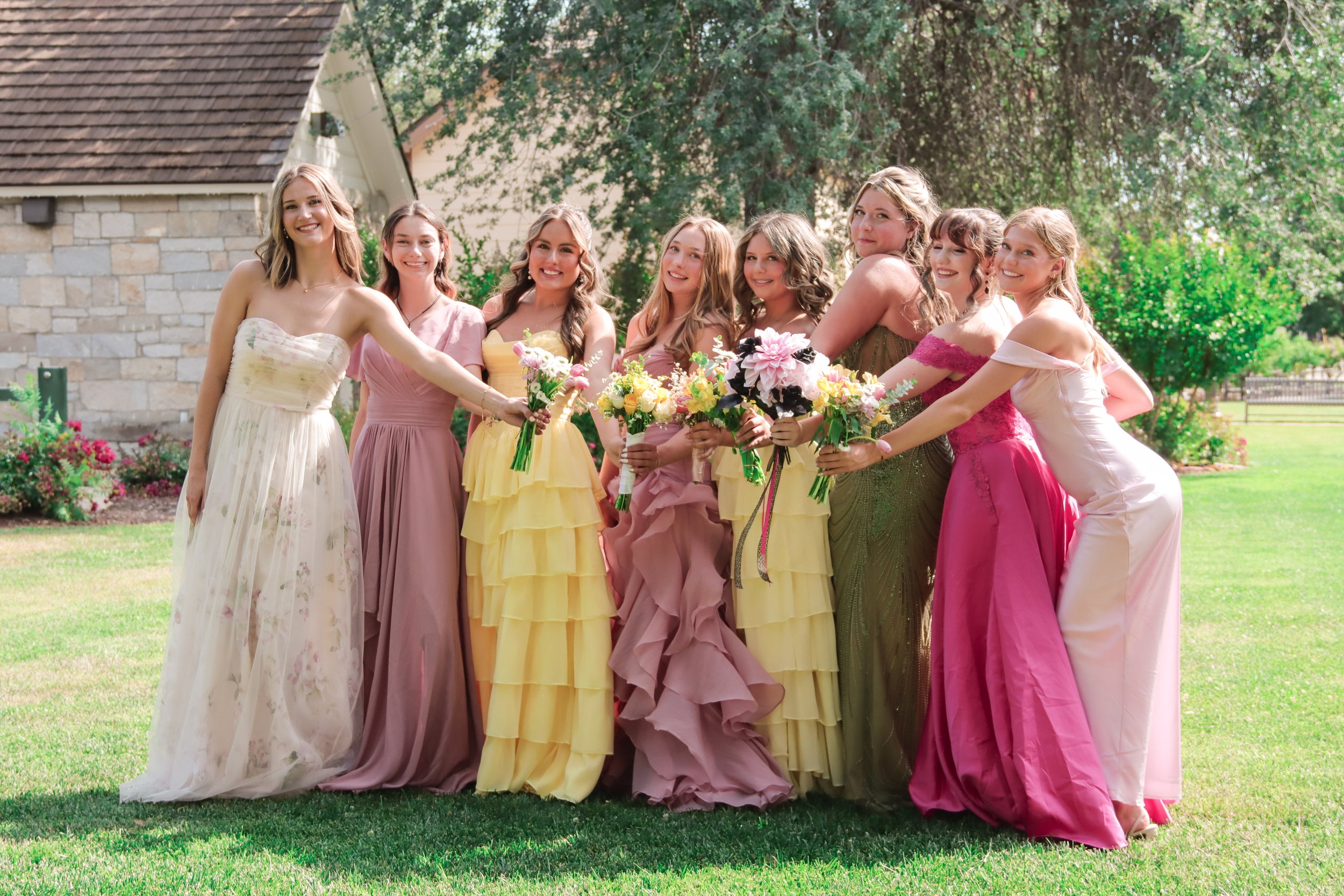 Group of women dressed in colorful formal gowns holding bouquets of flowers outdoors in a garden setting.