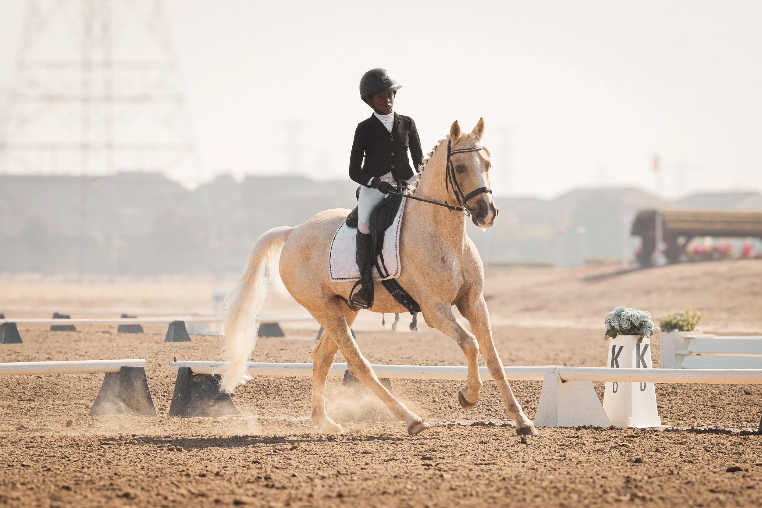 A young woman wearing formal riding attire, including a helmet, riding a light-colored horse in an outdoor competition arena with markers and a blurred background.