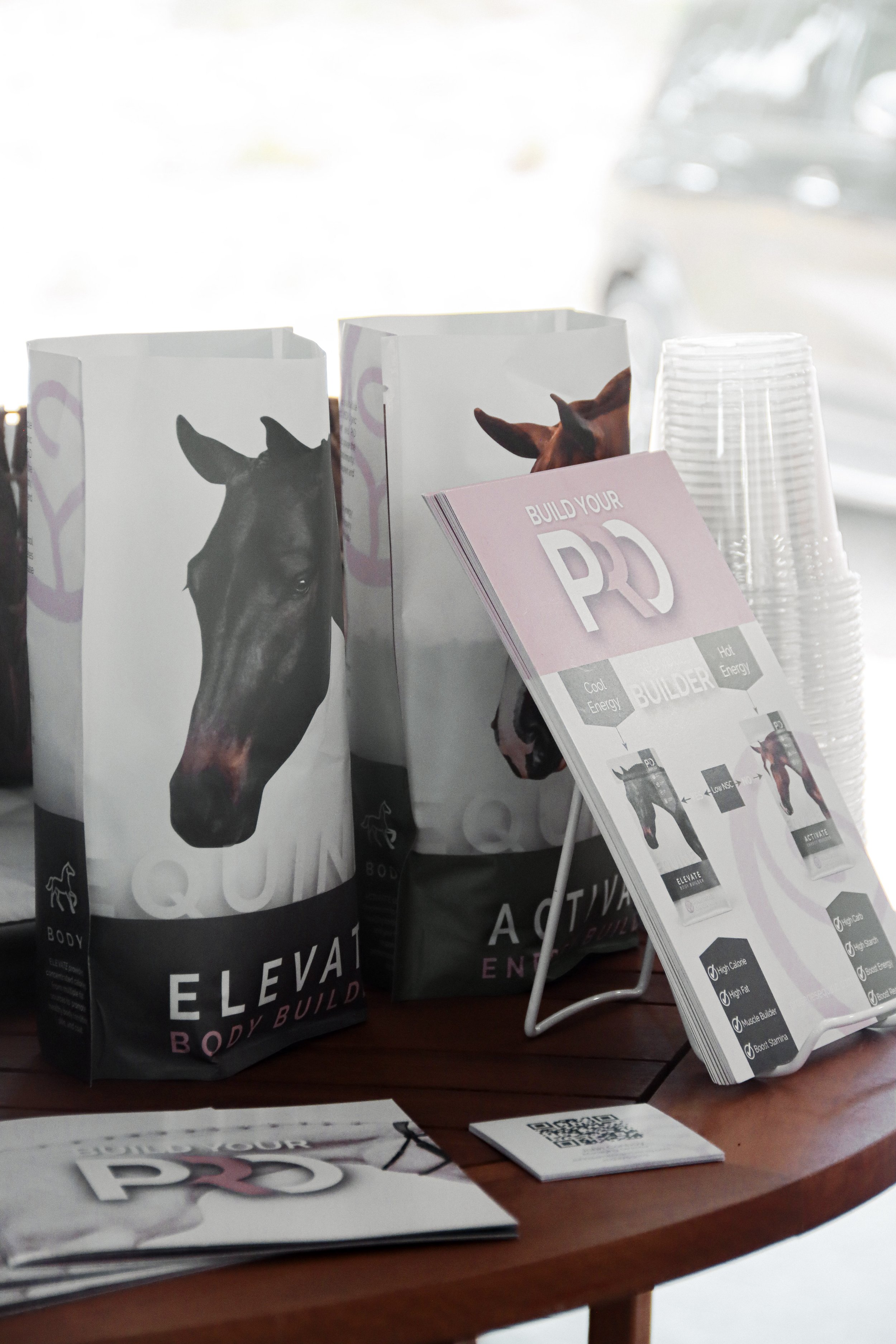 Display promoting an at-home body workout program called 'Elevate' with images of a horse on the packaging, on a table with informational brochures and stacked clear plastic cups.