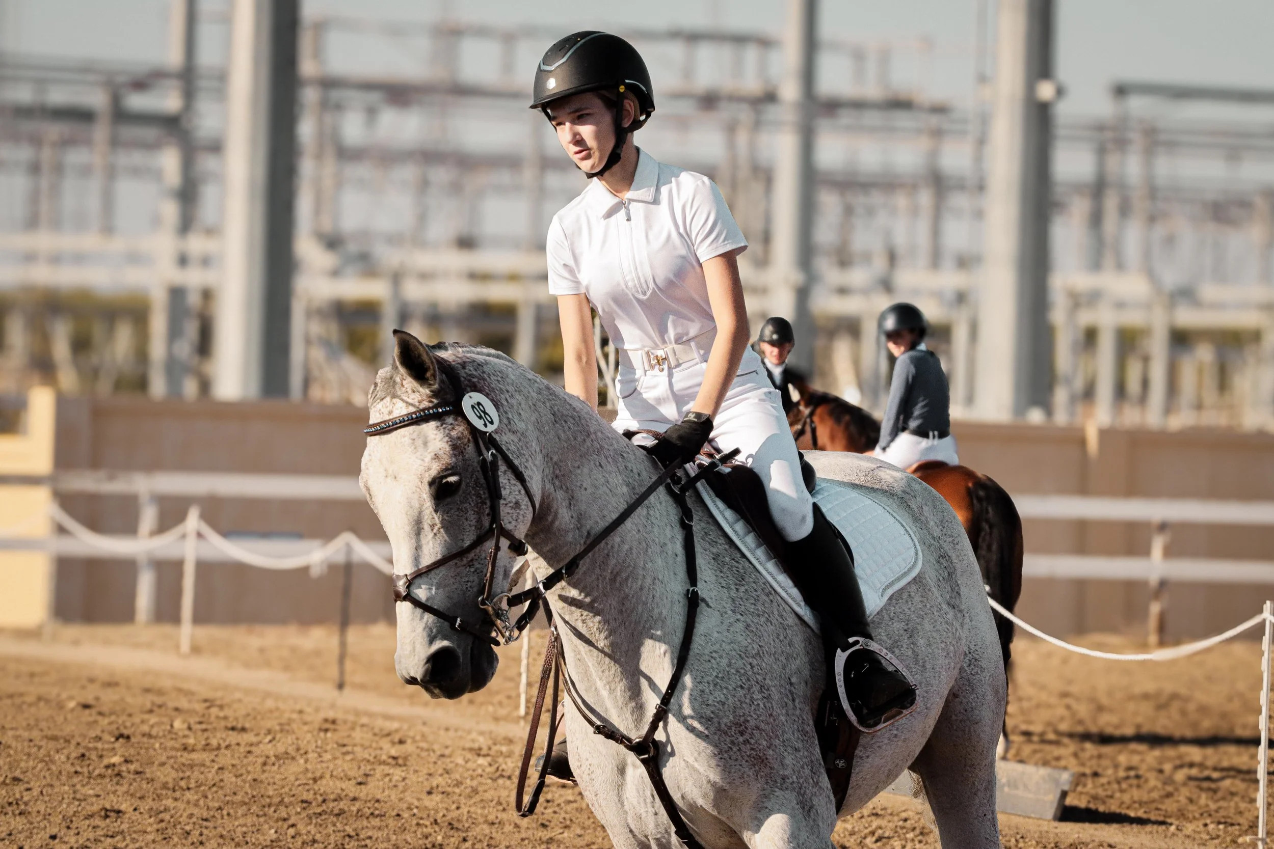 A woman wearing a white outfit and helmet riding a gray horse during a equestrian event on an outdoor dirt arena with other riders and spectators in the background.
