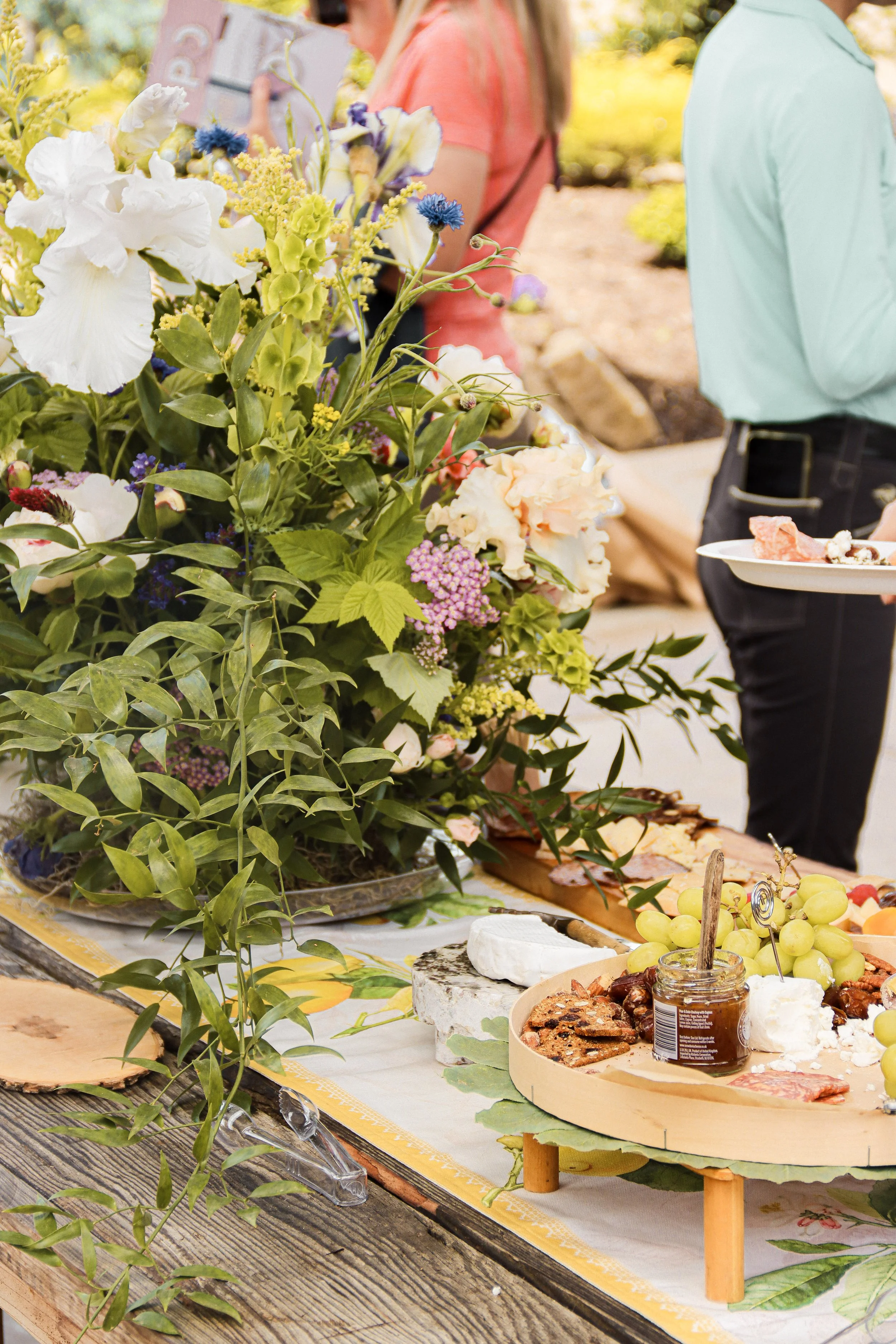 A table set with a variety of foods including grapes, cheese, crackers, and jam, decorated with a large bouquet of mixed flowers and greenery, outdoors with people in the background.