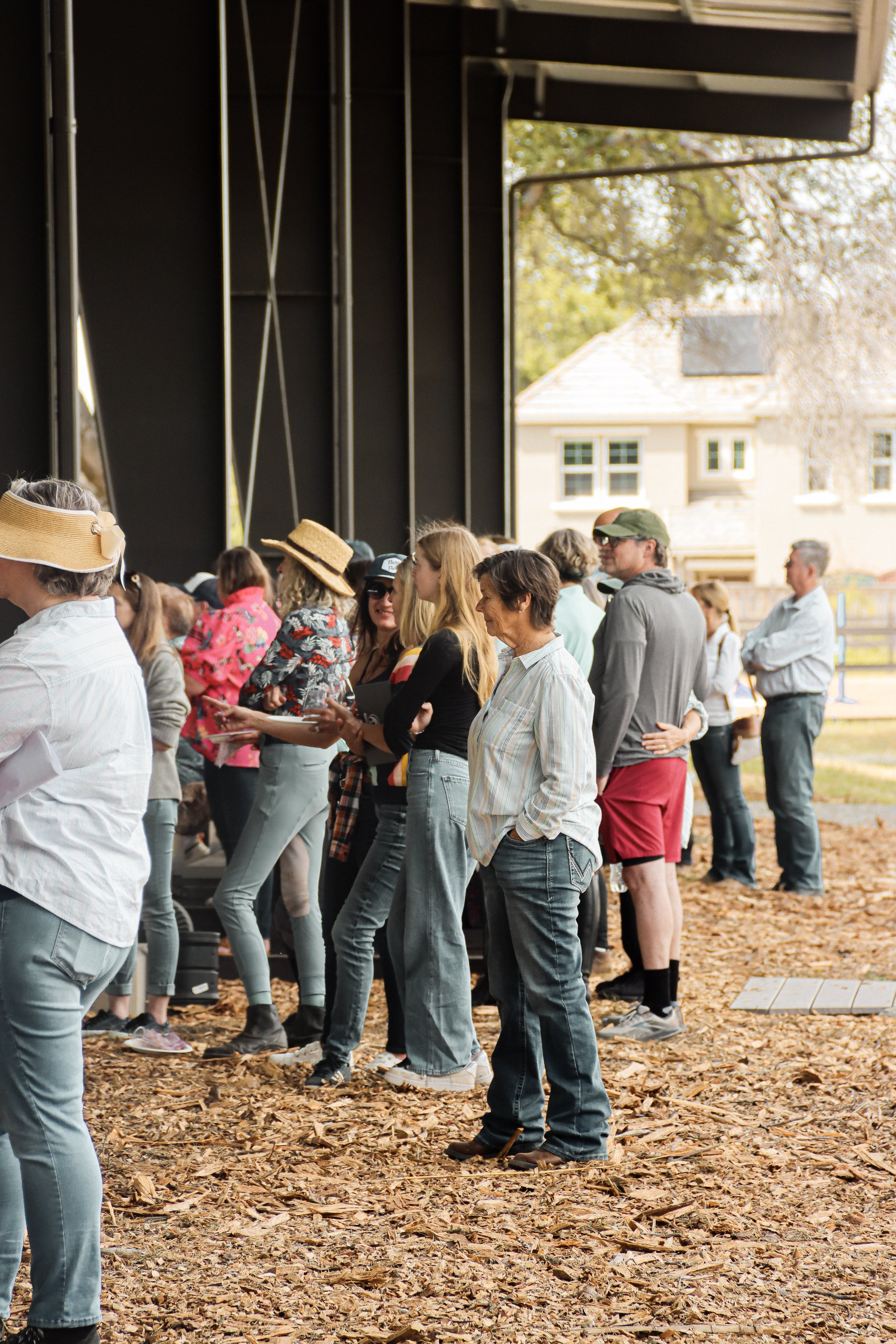 People standing in line outdoors on a leaf-covered ground, wearing casual clothing and hats, with a house and trees in the background.
