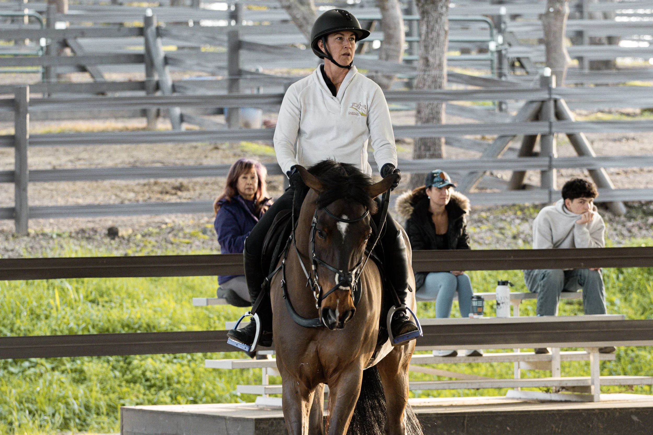 A woman wearing a white jacket and black helmet riding a brown horse at a race track with spectators sitting on benches in the background.