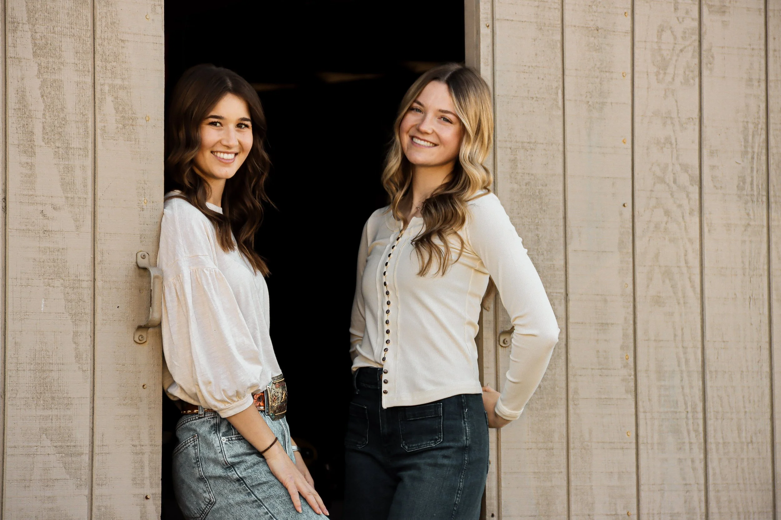 Two women with long hair standing outside a wooden building, smiling at the camera.