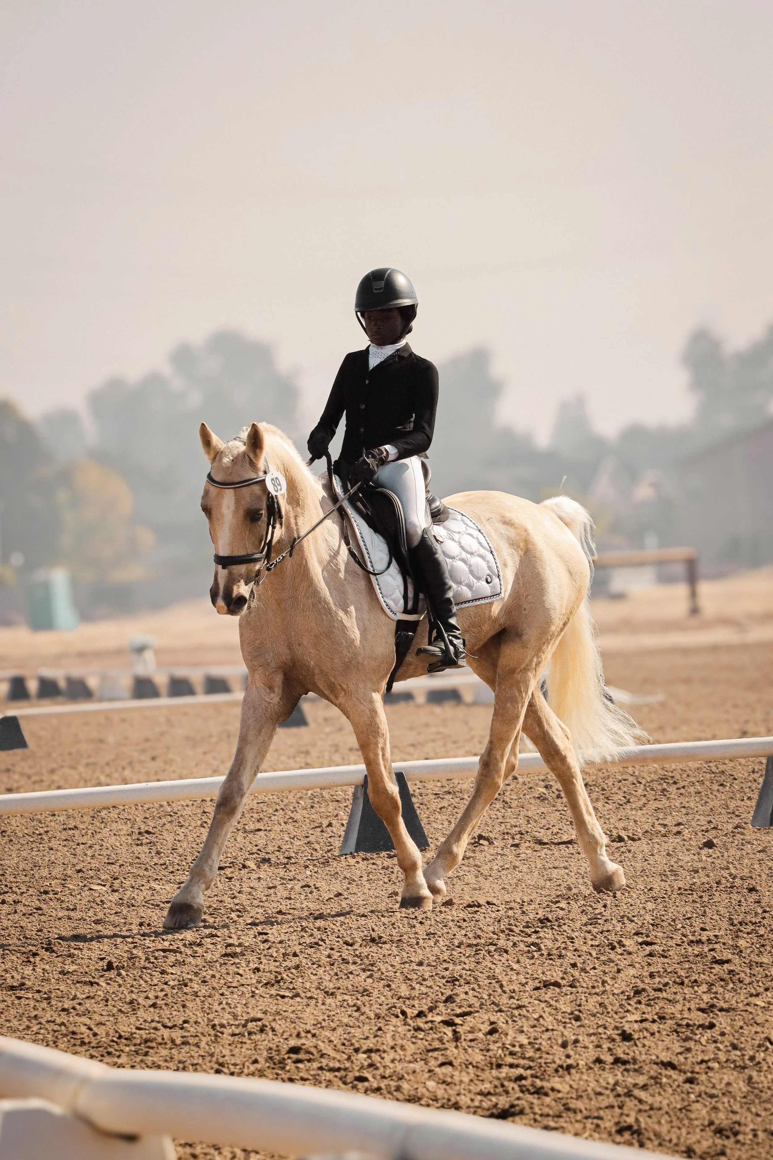 A person dressed in equestrian riding attire, including a helmet, riding a light-colored horse with a white saddle blanket, on a dirt riding arena with blurred trees and structures in the background.