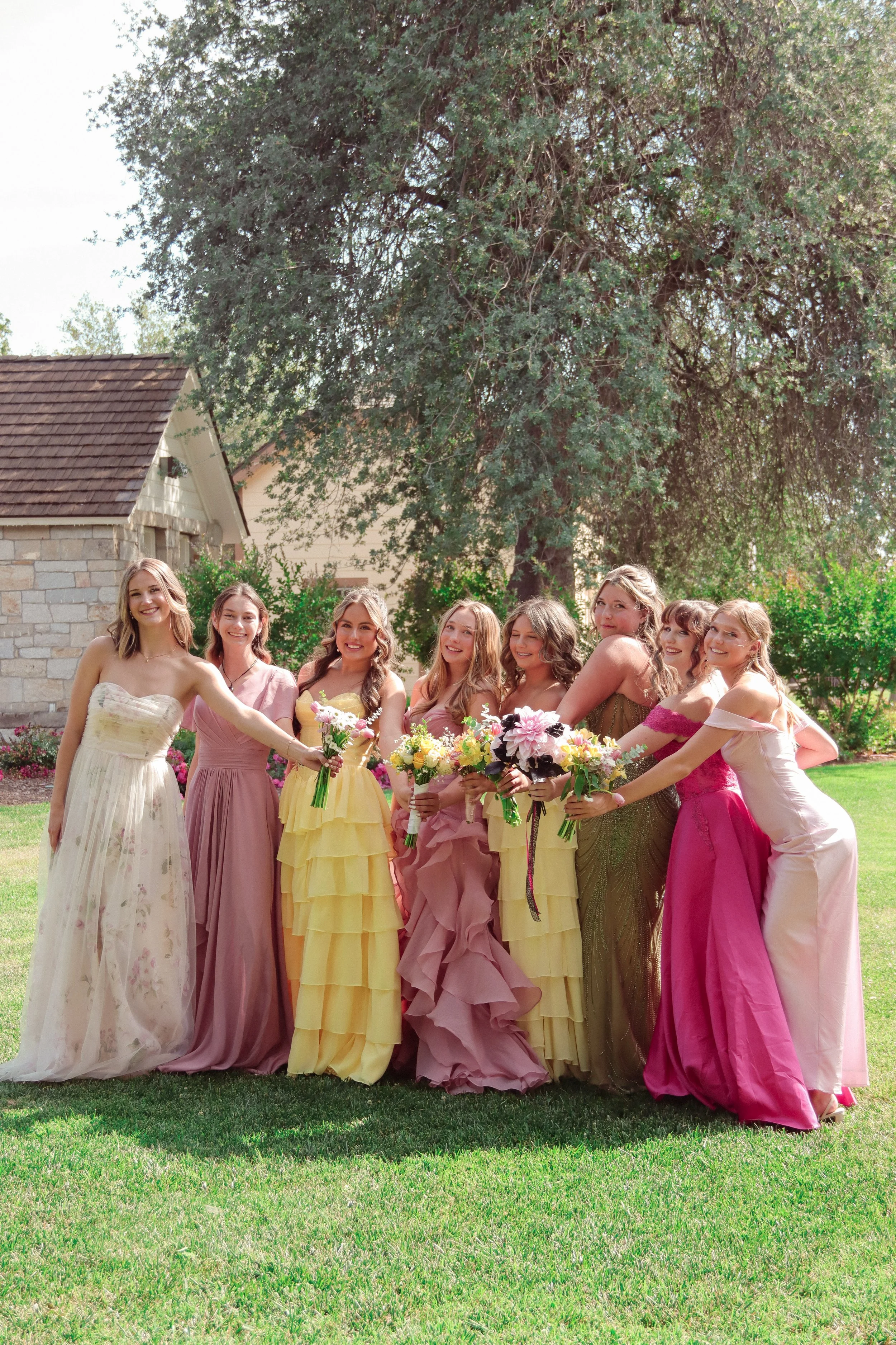 A group of women wearing colorful formal dresses standing outdoors in a garden with a large tree and a brick building in the background, holding bouquets of flowers.