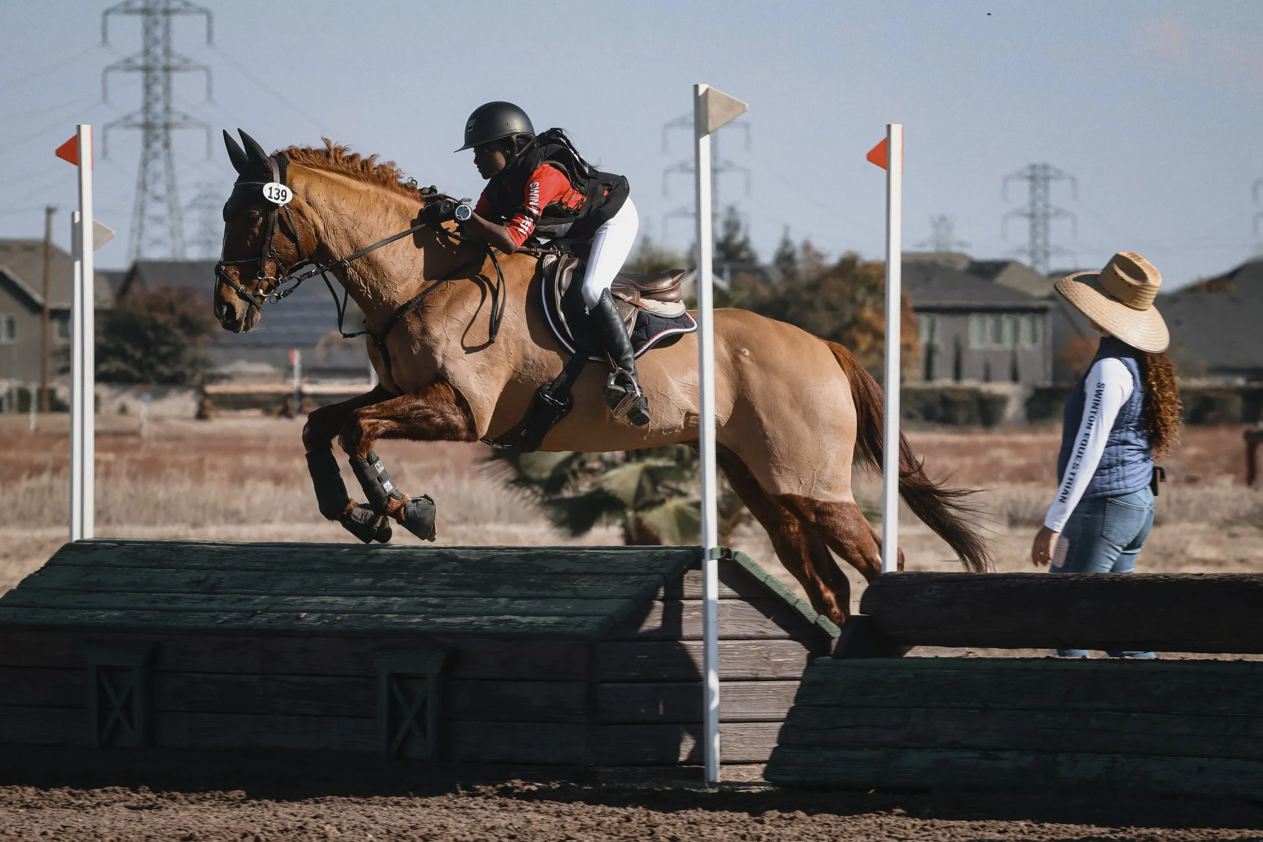 A rider on a chestnut horse is jumping over an obstacle during a horseback riding competition. A woman wearing a large straw hat is standing nearby, watching the event with houses and power lines in the background.