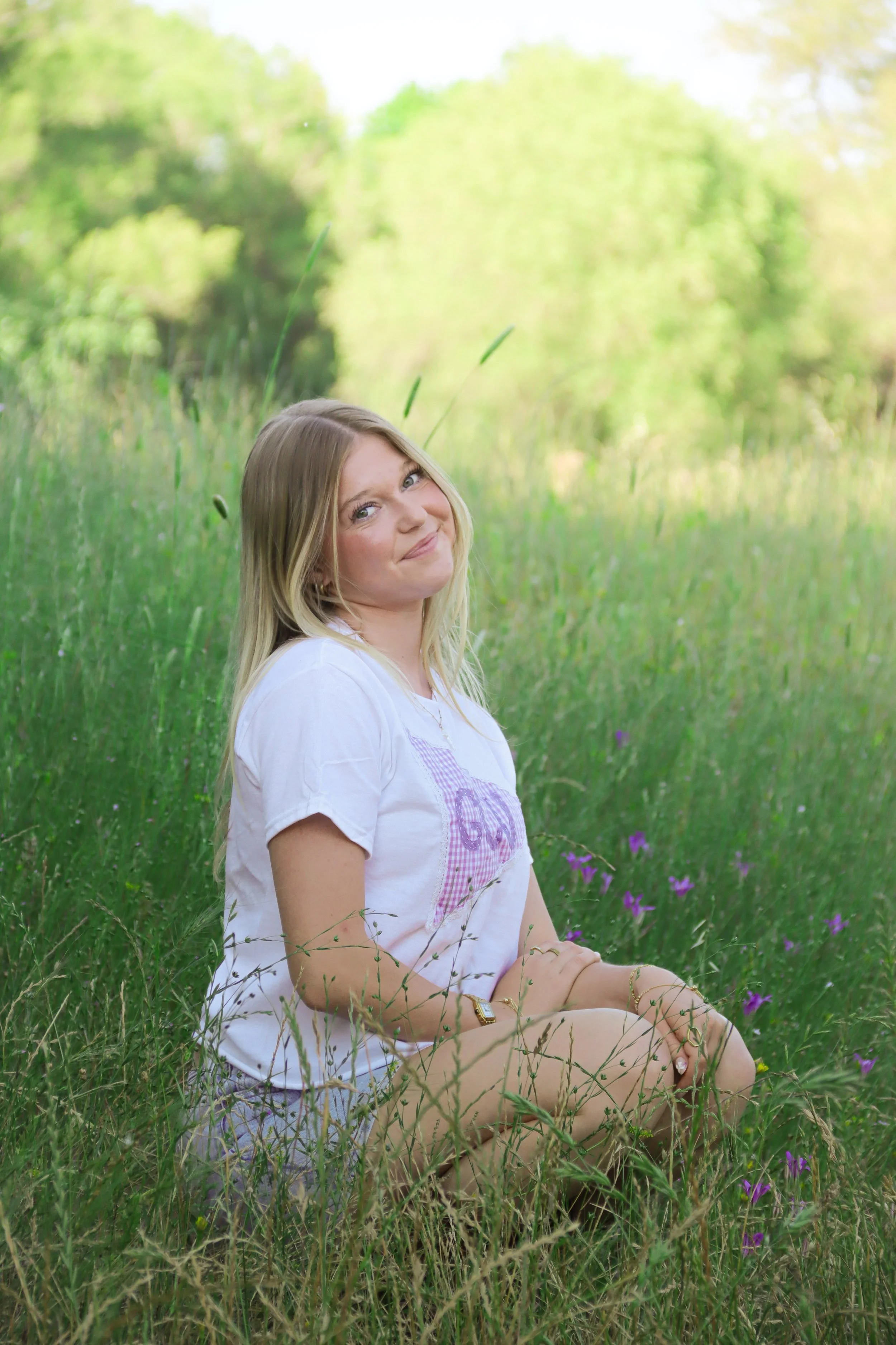 A young woman with blonde hair sitting in tall grass with purple flowers in a lush green field, smiling at the camera.