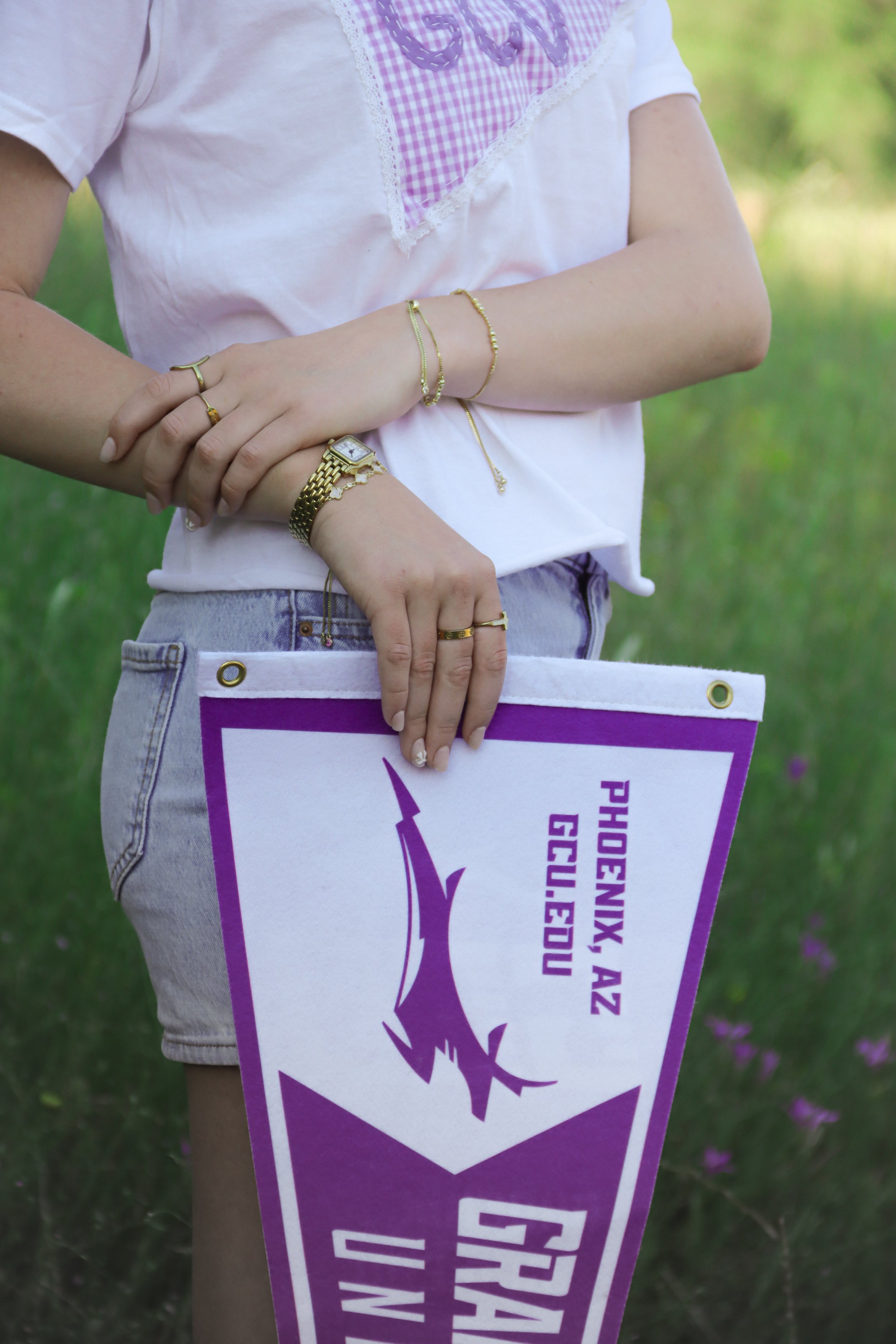 Person holding a purple and white sign outdoors, with bracelets, rings, and a watch on their hands, wearing a white top with a pink gingham pattern.