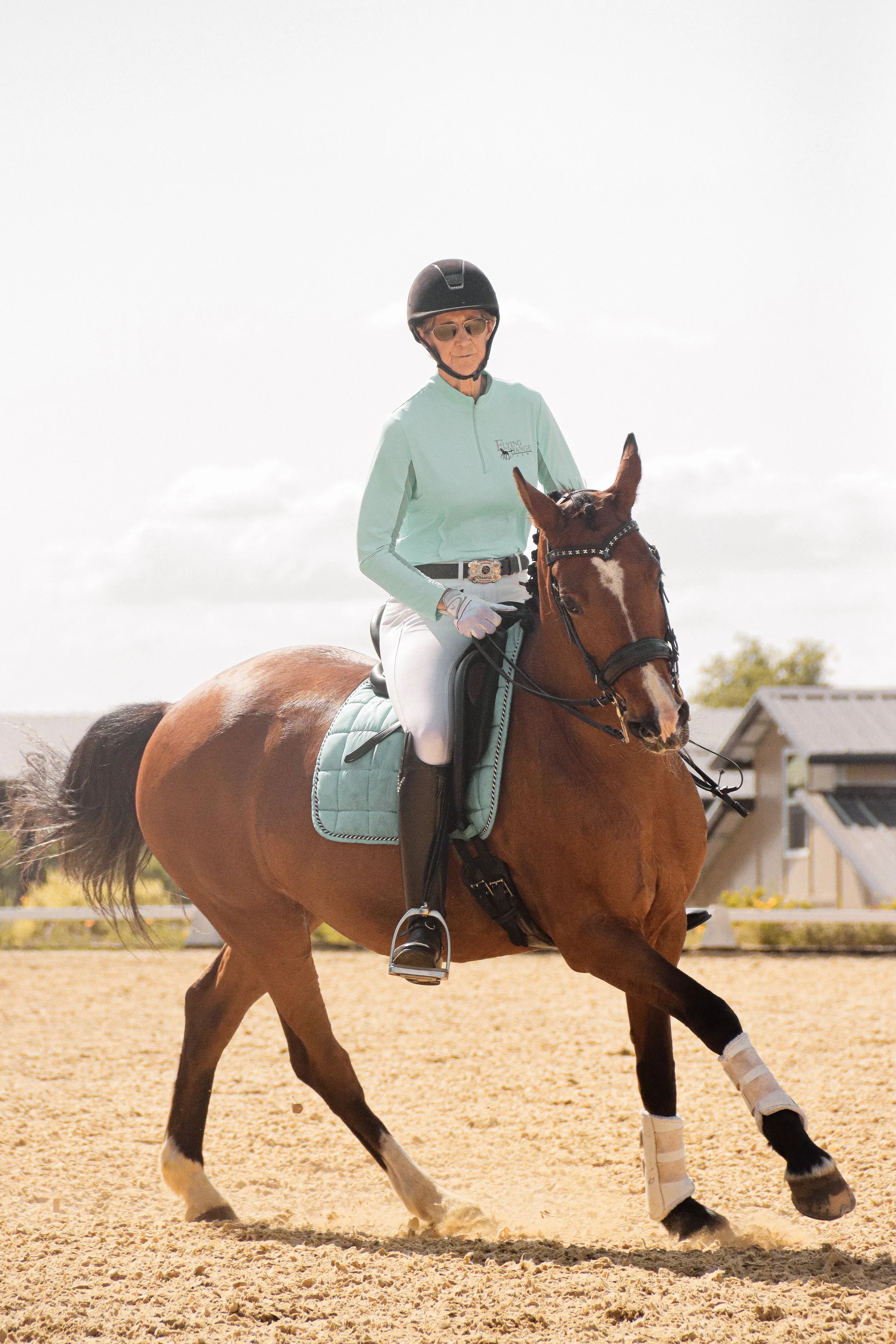 A woman in riding gear, wearing sunglasses and a helmet, riding a brown horse with white markings on its face and legs in an outdoor riding arena with a sandy surface.