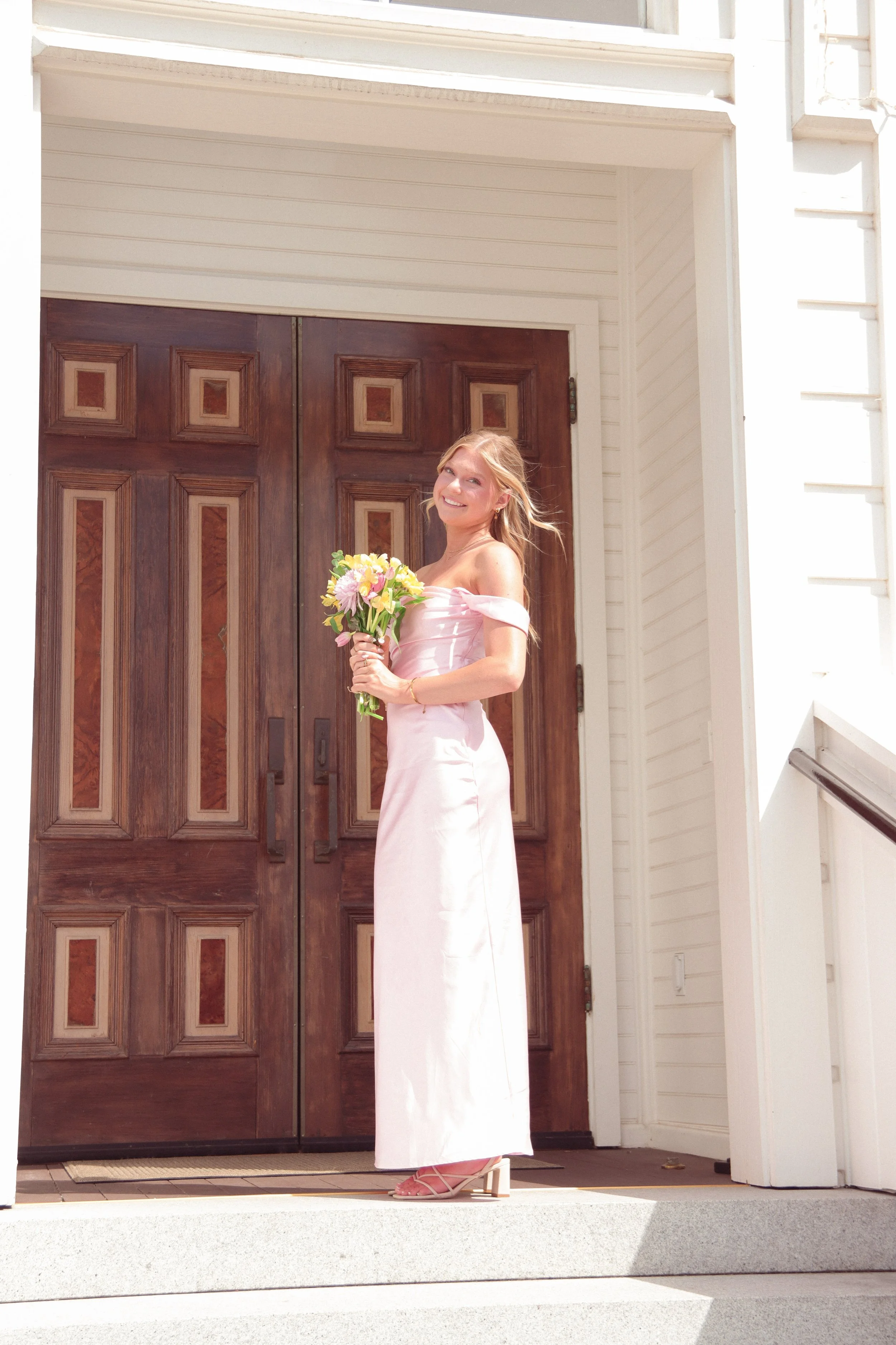 A woman in a long pink dress holding a bouquet of flowers standing in front of a wooden door on the steps outside a house.
