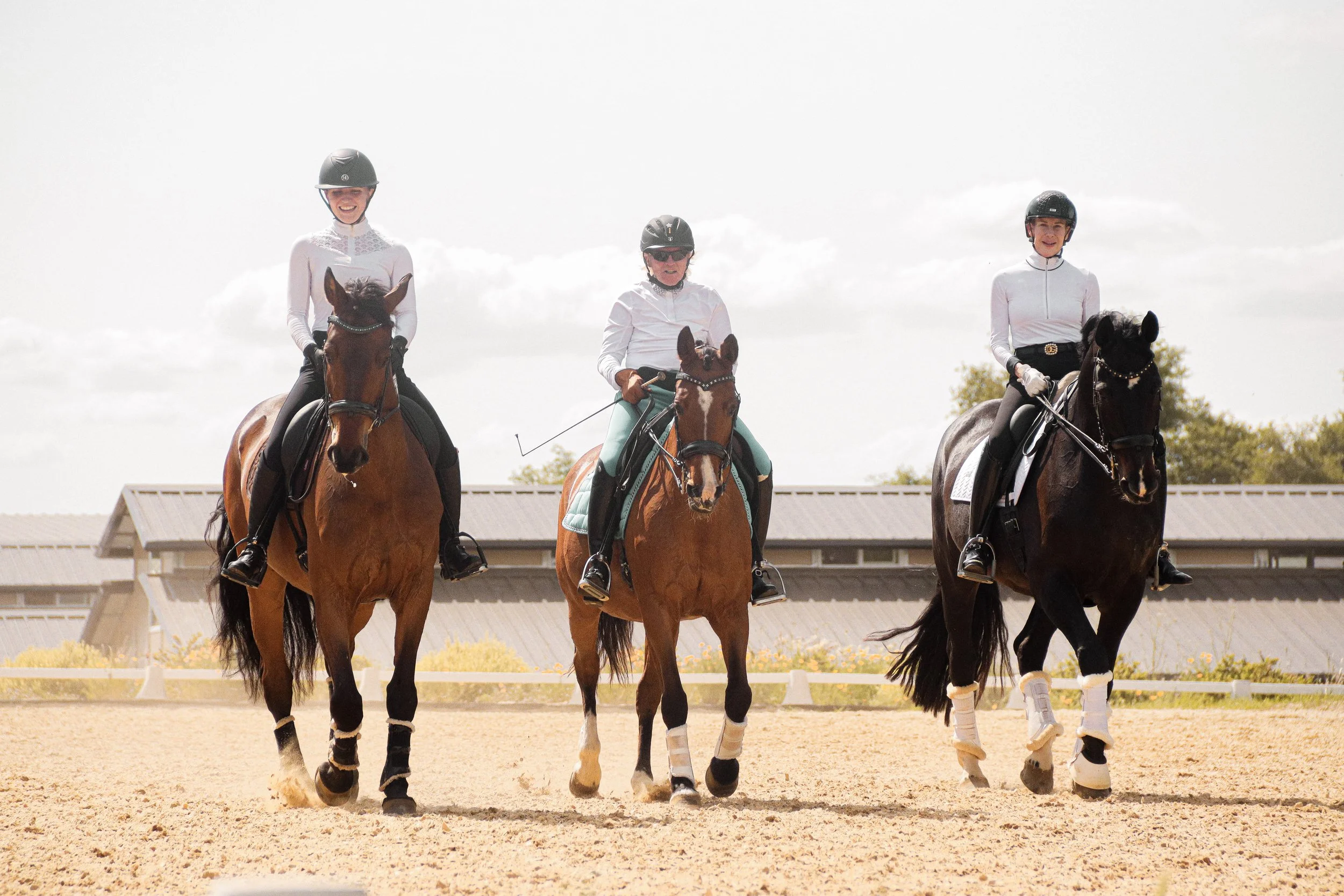 Three people riding horses on a sandy equestrian arena, wearing helmets, with a building and trees in the background.