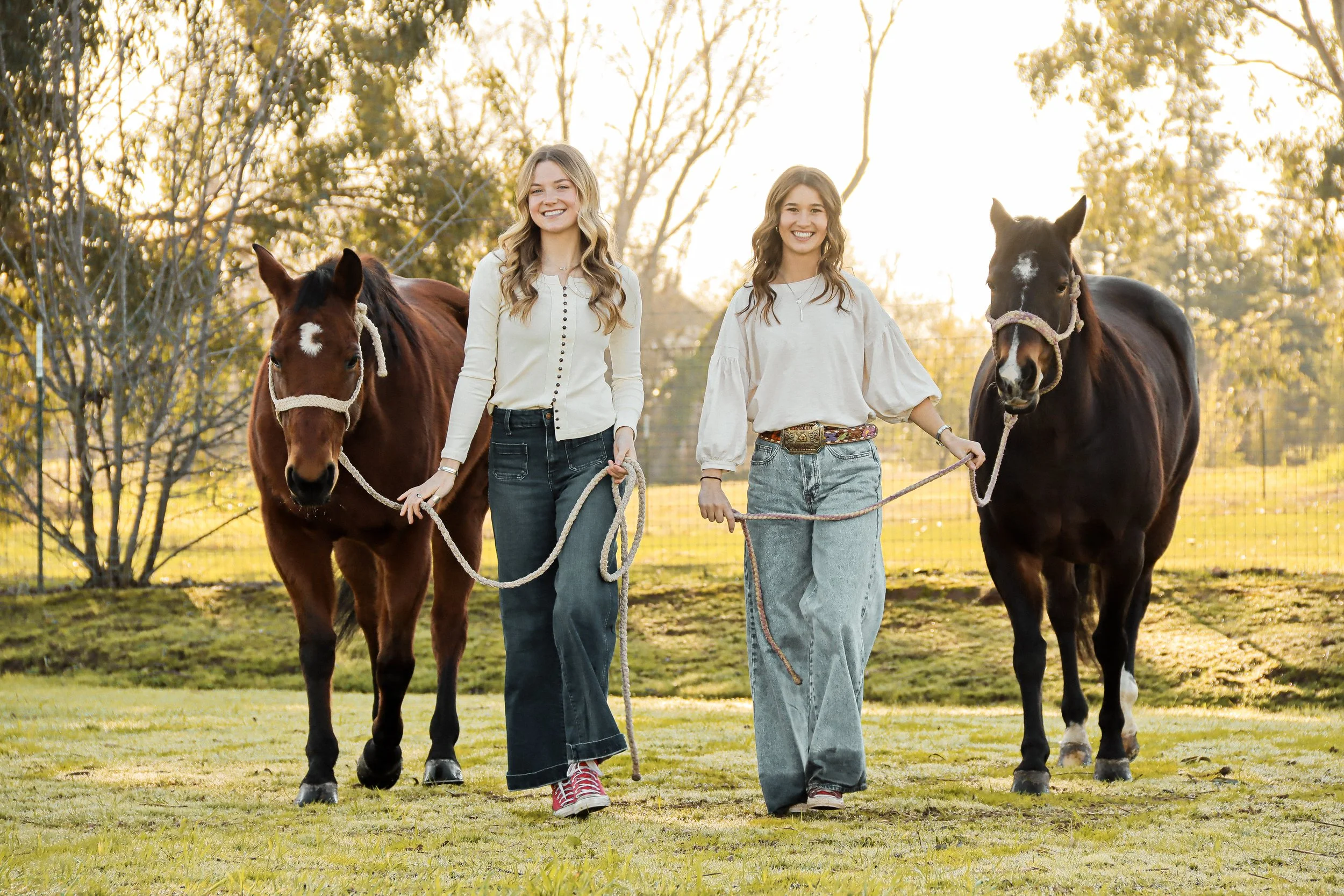 Two young women walking with ropes each holding a horse leads in a grassy field at sunset, trees in the background.