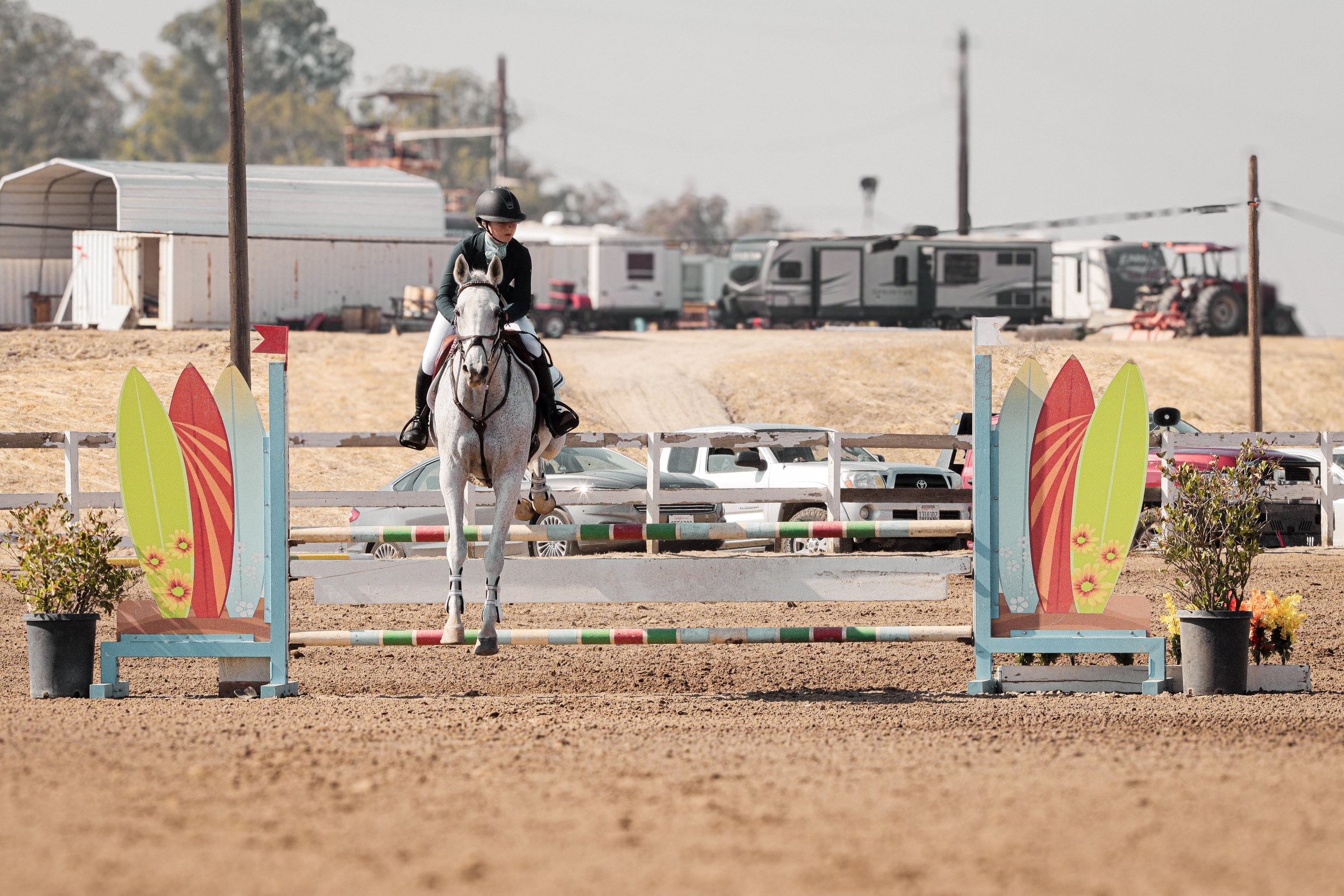 A female horseback rider jumps over a colorful obstacle on a sandy outdoor riding arena, with trailers and farm equipment in the background.
