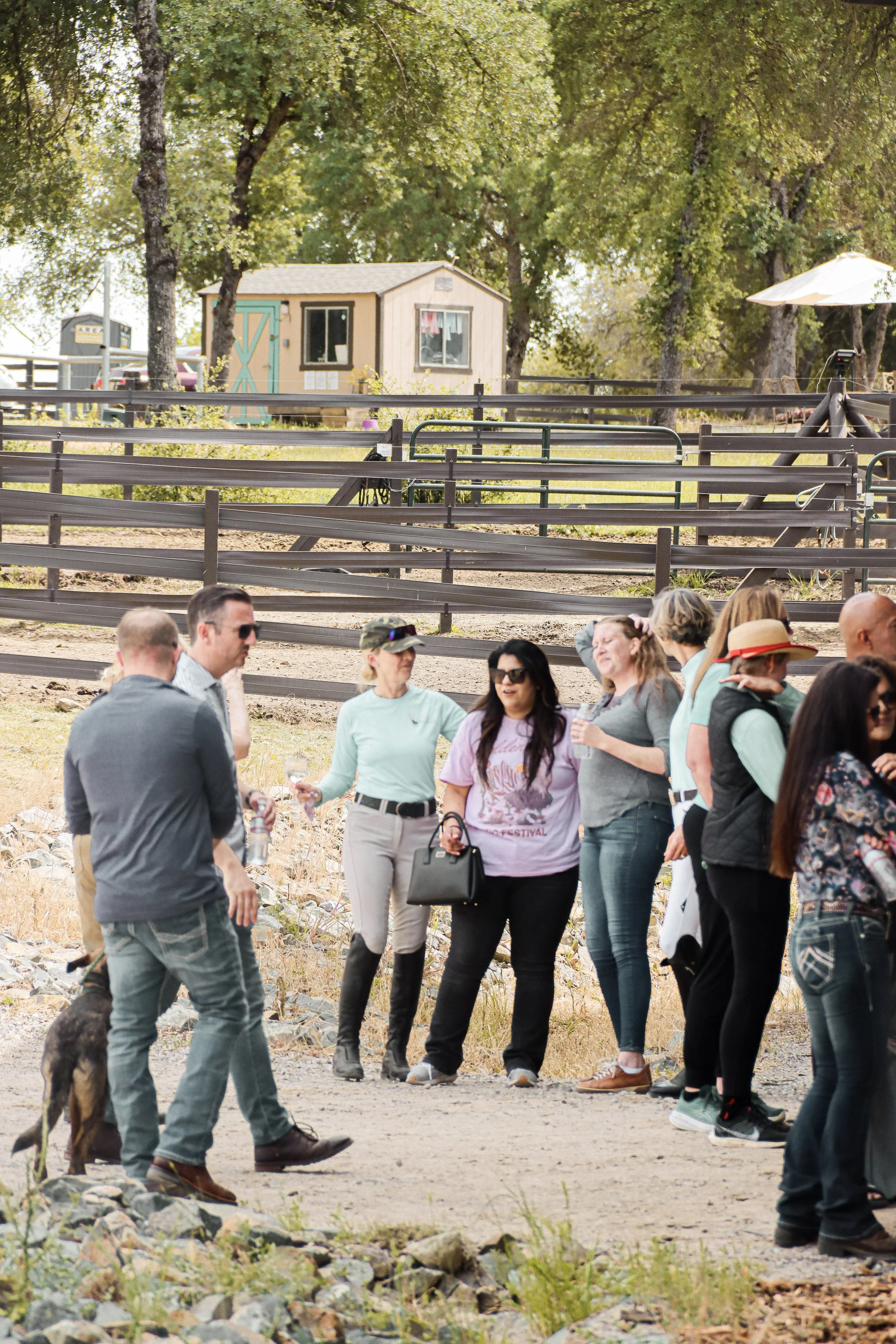 Group of people socializing outdoors in a rural setting with trees, a small shed, and a wooden fence in the background.