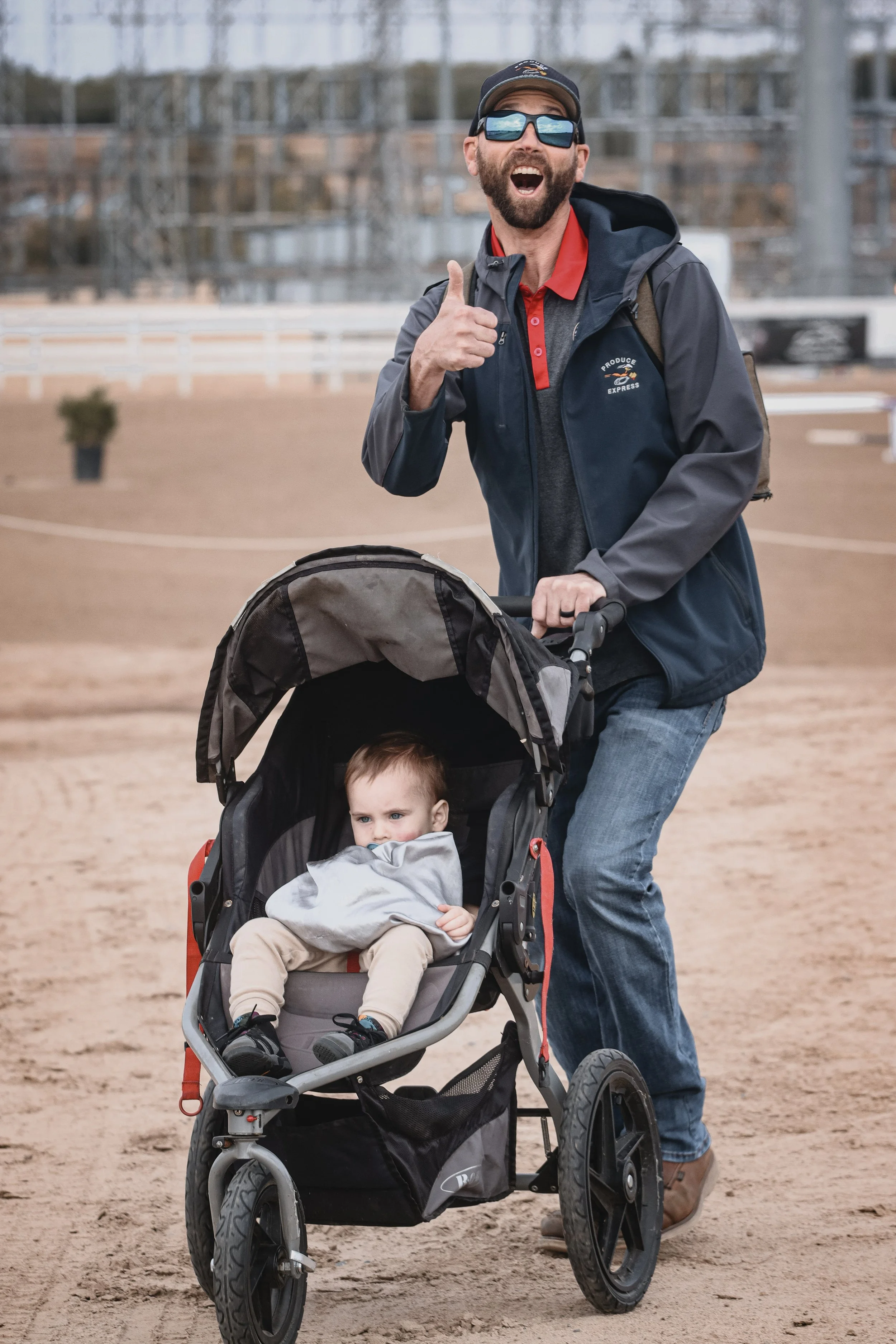 A man with a beard, dark sunglasses, and a black cap is riding an electric stroller with a young child sitting inside. The man is giving a thumbs-up and smiling widely, wearing a gray and navy blue jacket over a red polo shirt. The background shows a