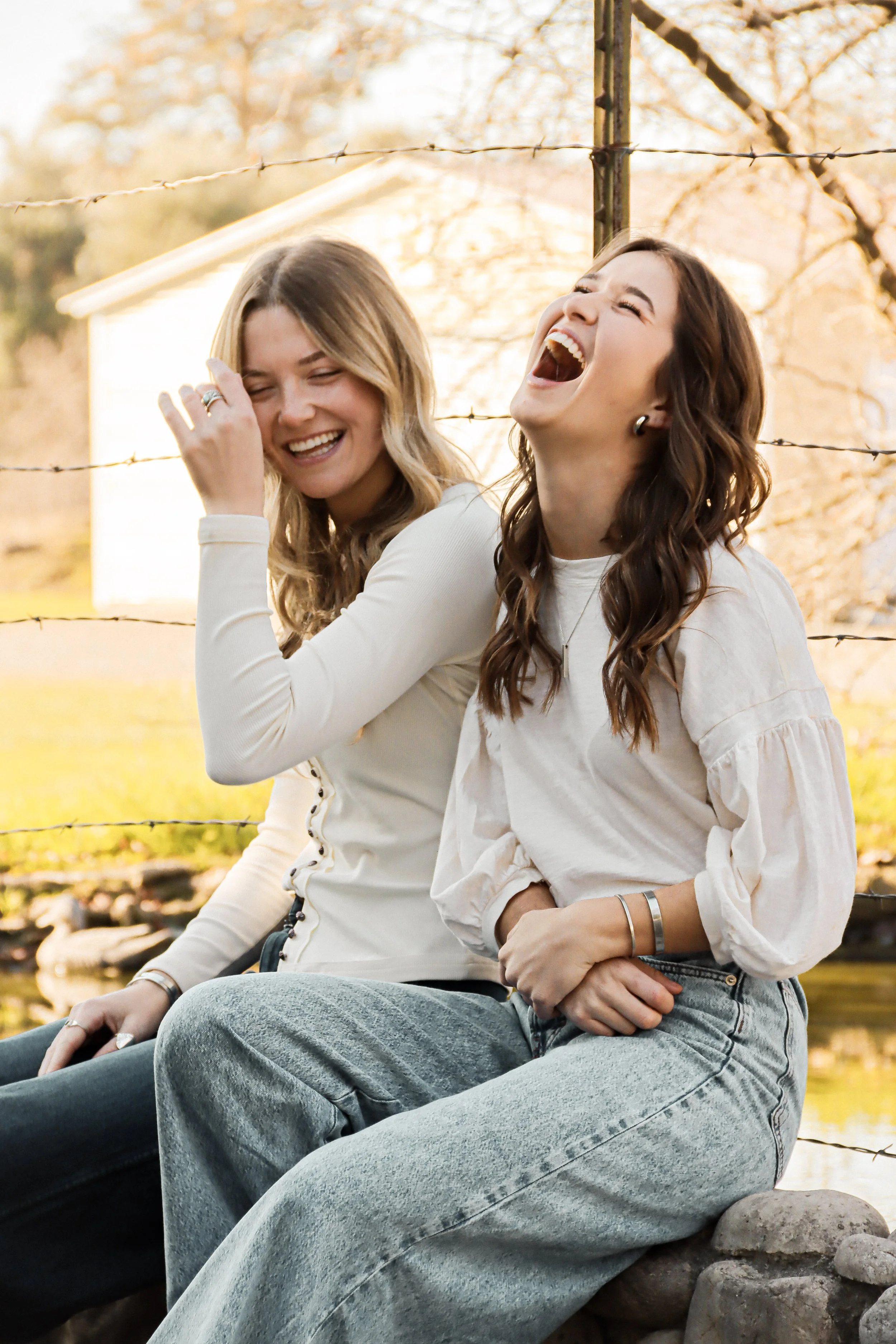 Two women sitting outdoors near a barbed wire fence, laughing and enjoying each other's company during autumn with trees and a building in the background.