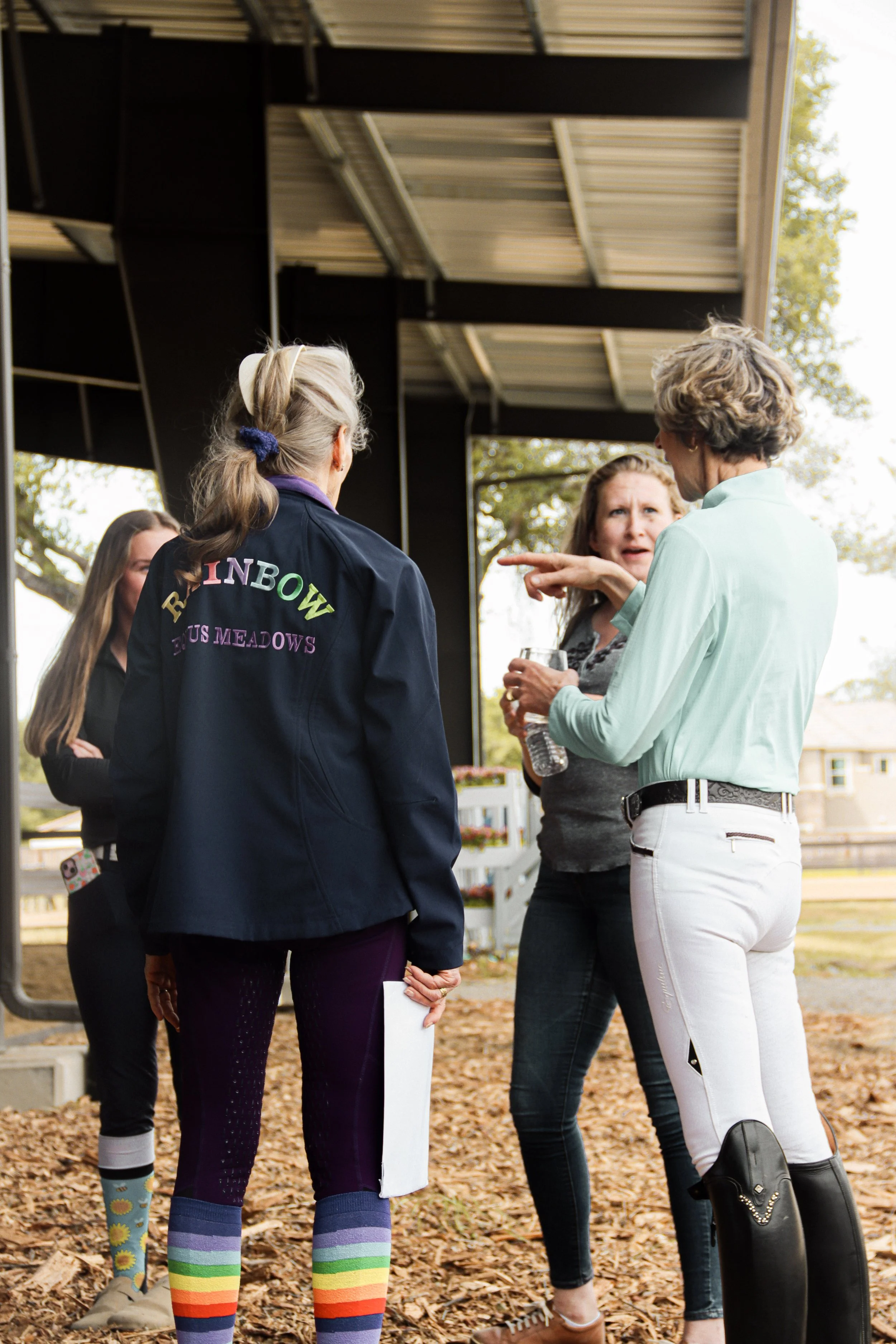 Four women standing outdoors on a fall day, engaged in conversation. One woman, wearing a mint green blouse and white pants, is gesturing as she speaks. Another woman, wearing a dark jacket with colorful embroidered words, holds a water bottle. The t