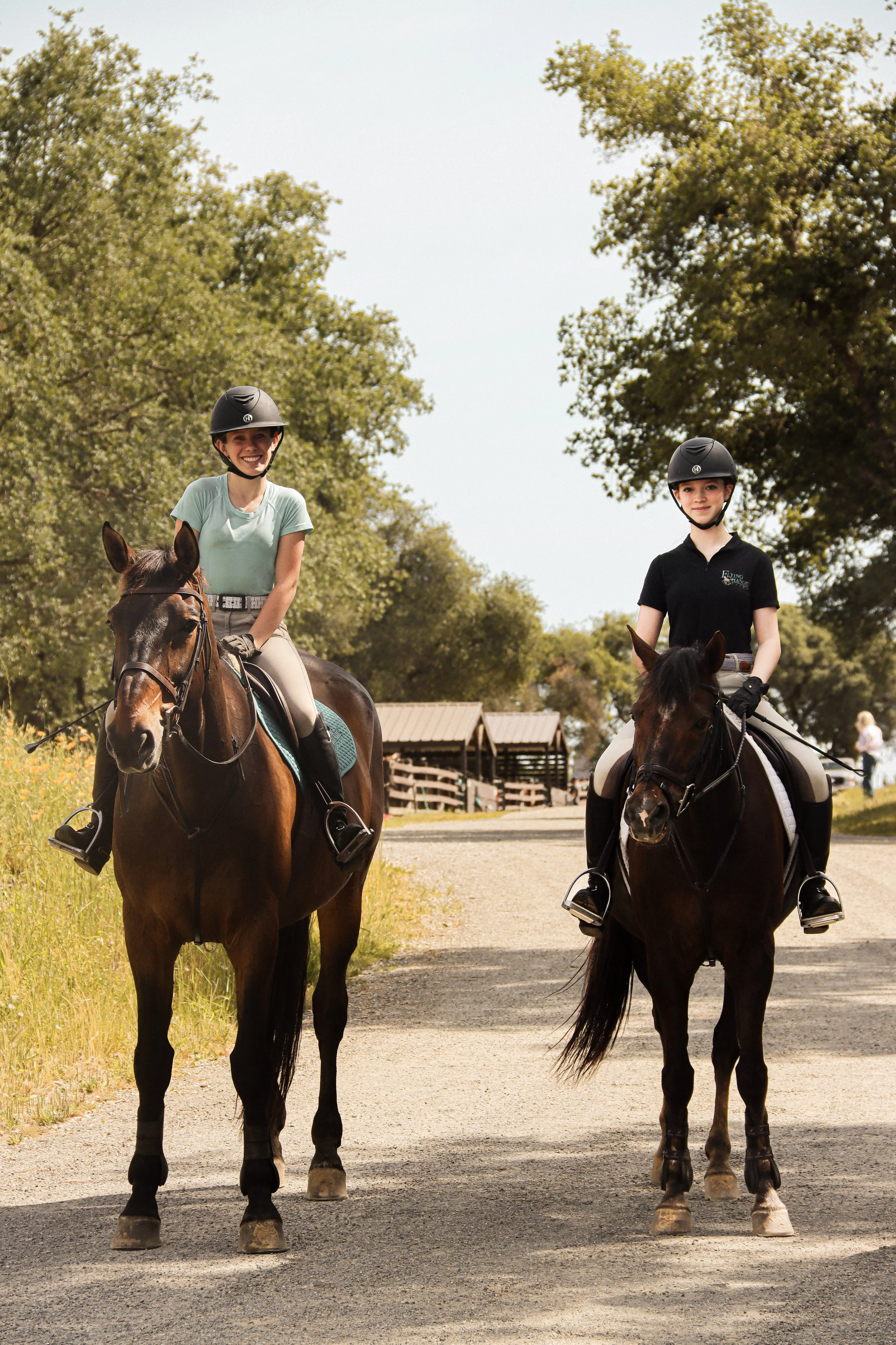Two young women riding horses outdoors on a sunny day, wearing helmets and riding gear, with trees and a farm building in the background.