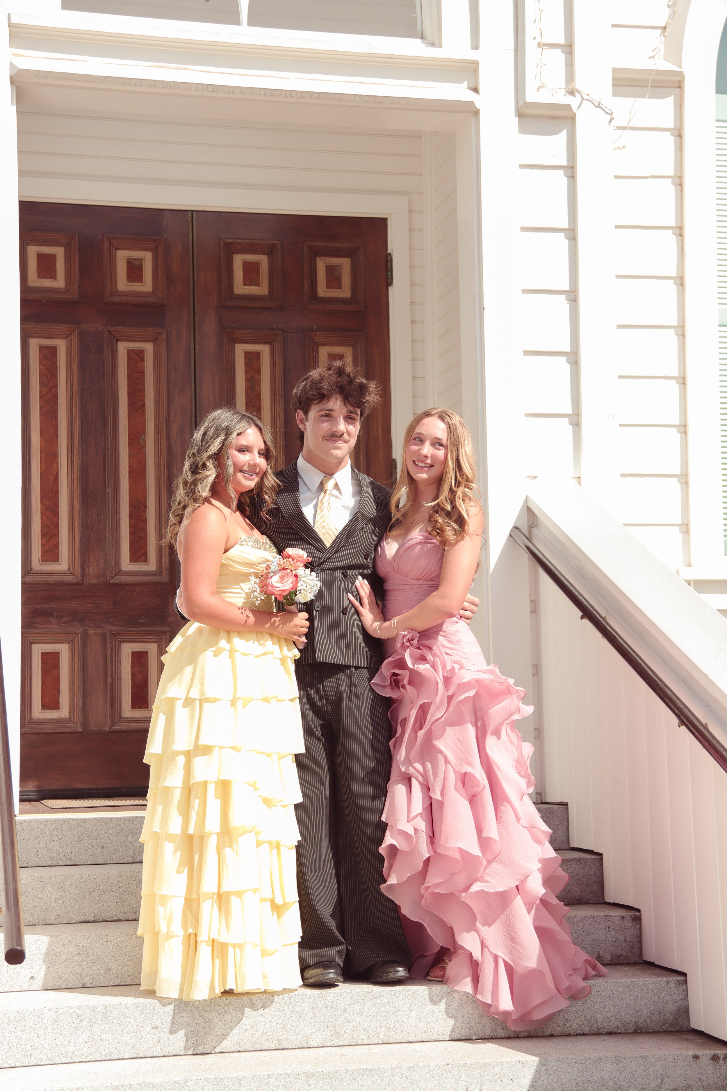 Three people standing on stairs in front of a wooden door. The two women are wearing formal dresses, one in yellow and the other in pink, and the man is wearing a black suit with a tie. The woman in pink is holding a bouquet of flowers.