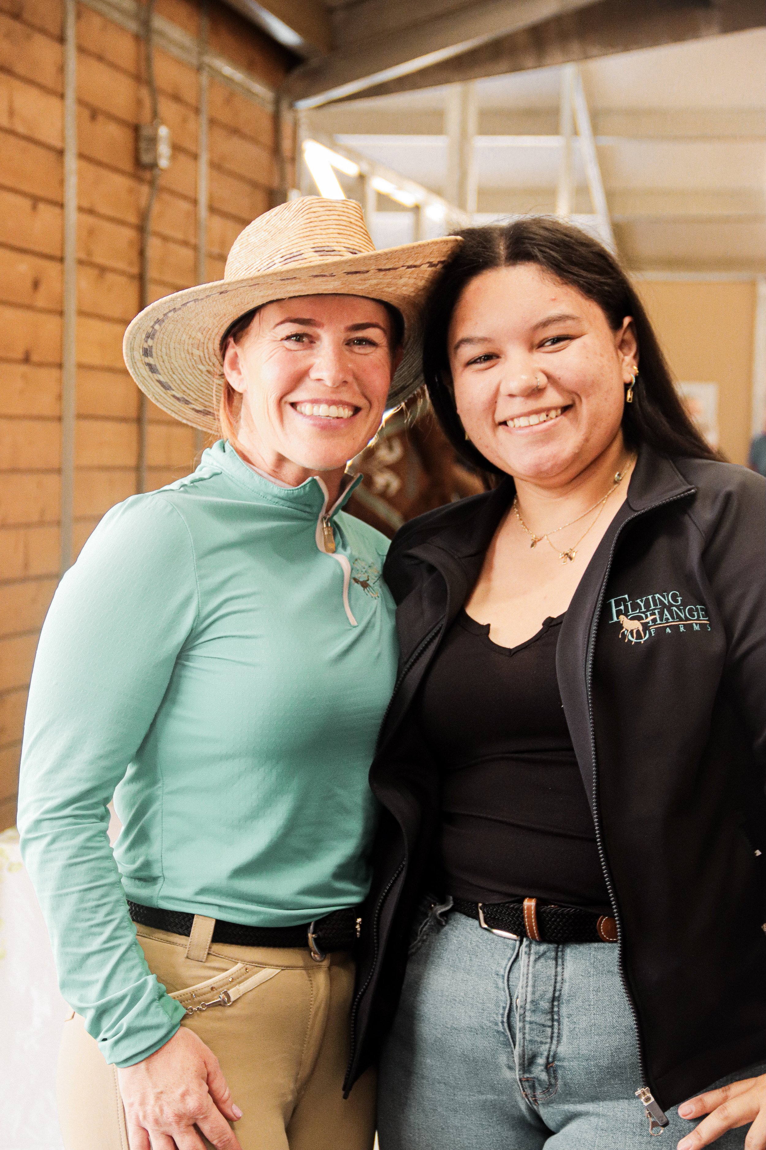 Two women smiling, standing close together indoors. One wears a large straw hat and a light green zip-up jacket, the other wears a black jacket with a logo that says "Flying Change Farm" and has a small mustache. Both women are dressed casually.