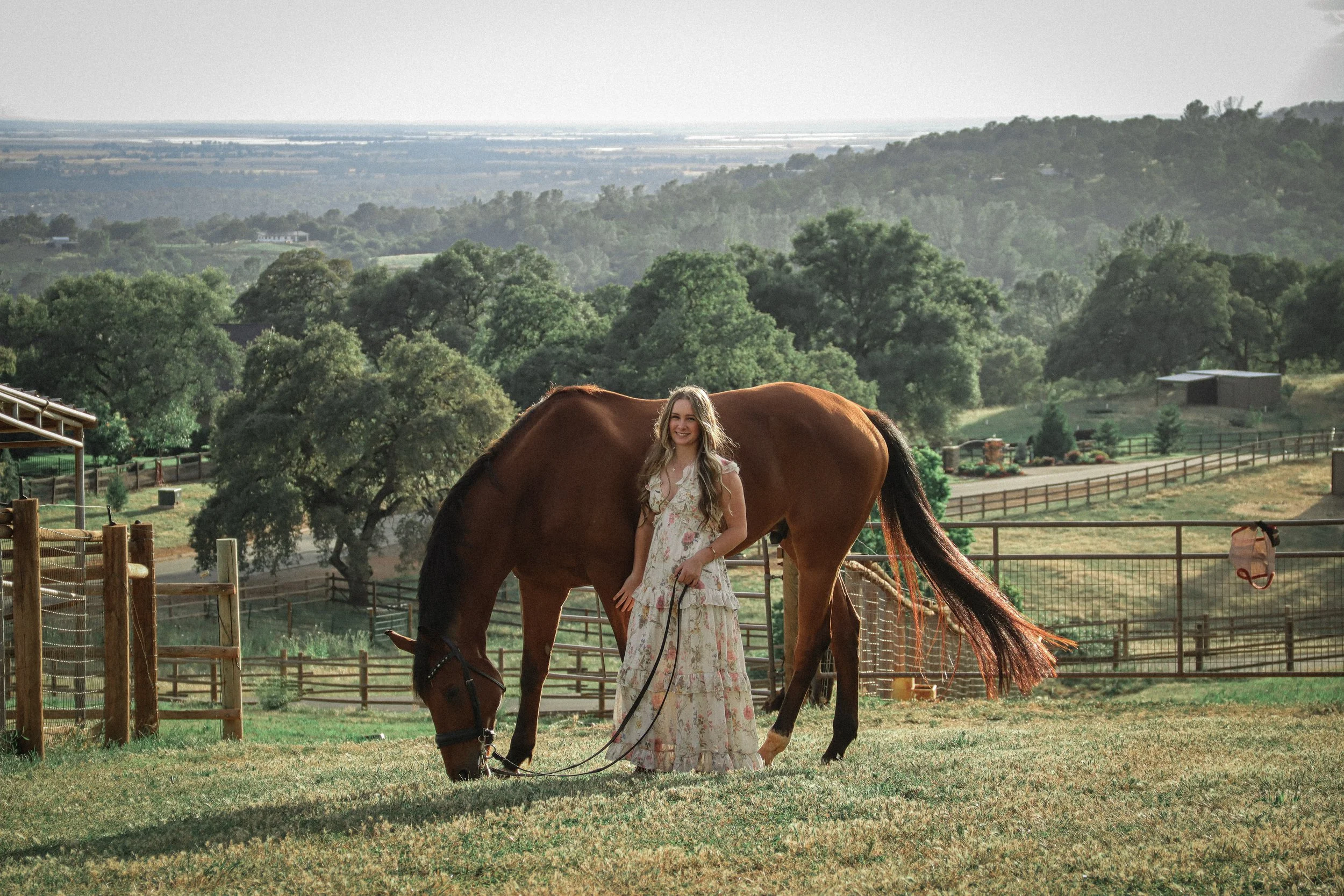 A young woman with long blonde hair in a floral dress standing next to a brown horse on a farm, with trees, fencing, and distant hills in the background.