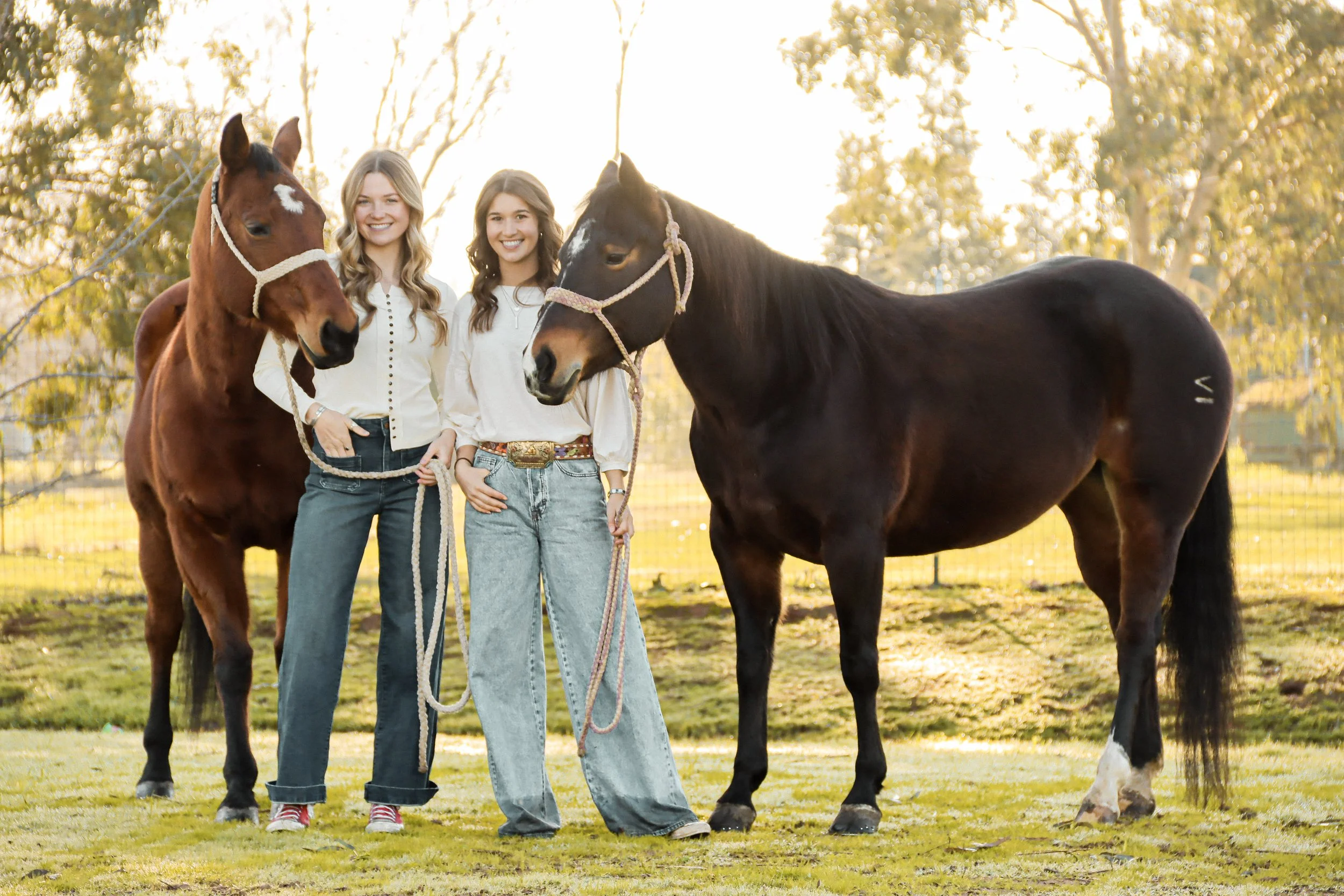 Two women standing outdoors in a field during golden hour, each holding a horse's lead rope, with trees and sunlight in the background.