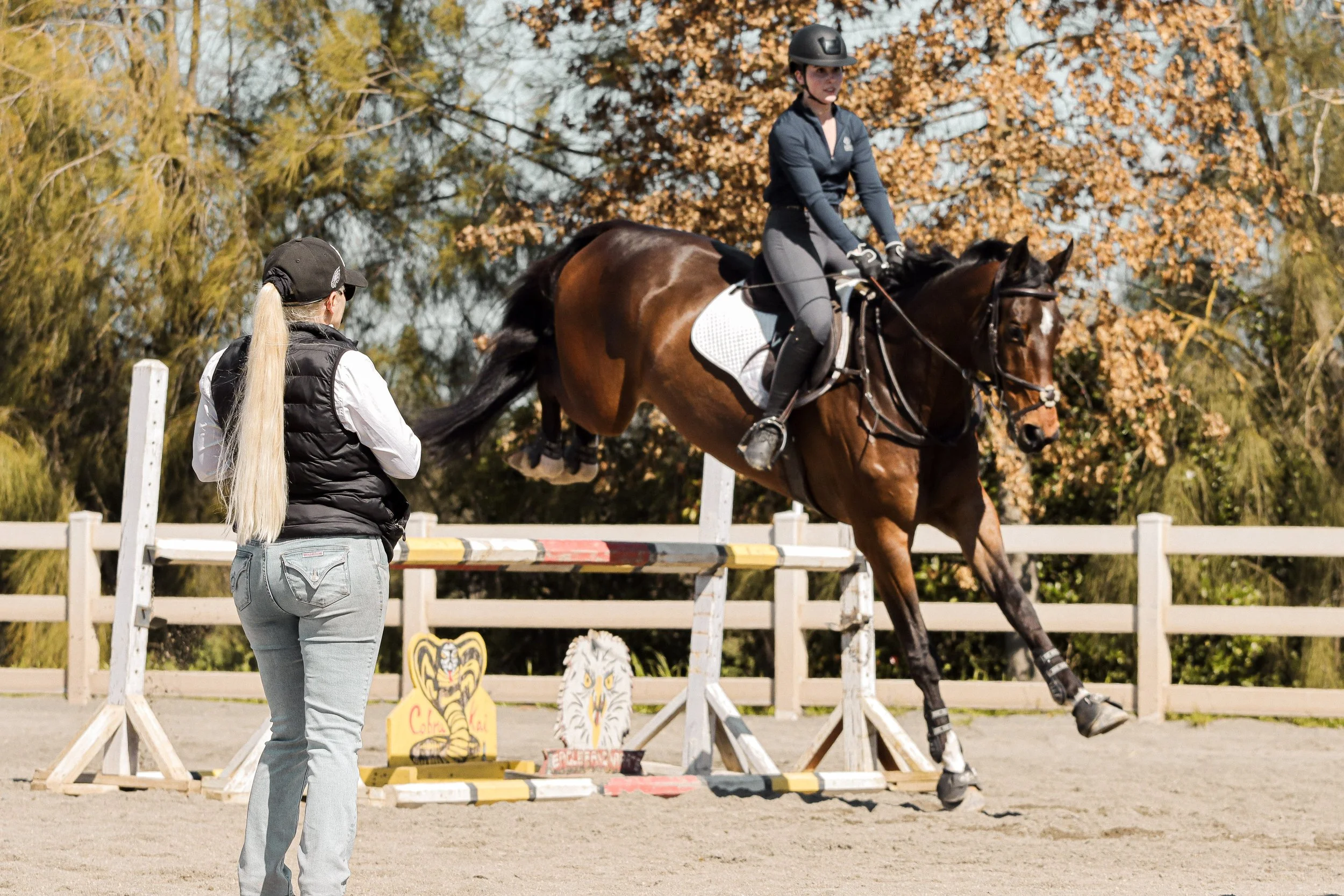 A woman standing near a jumping horse during an equestrian event with fall-colored trees in the background.