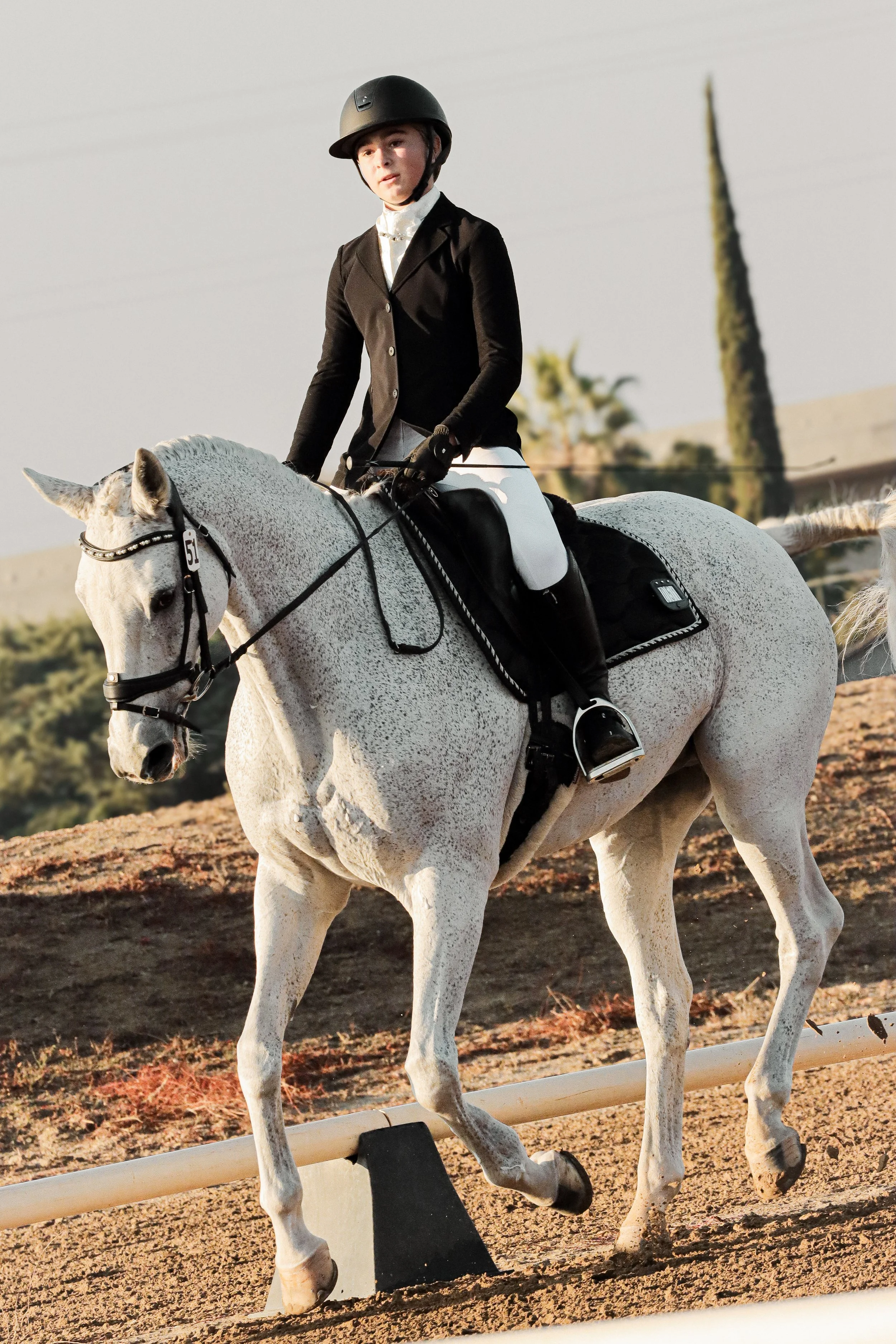 A person riding a white horse during a horseback riding competition, wearing a black helmet and riding attire, with a scenic outdoor background.