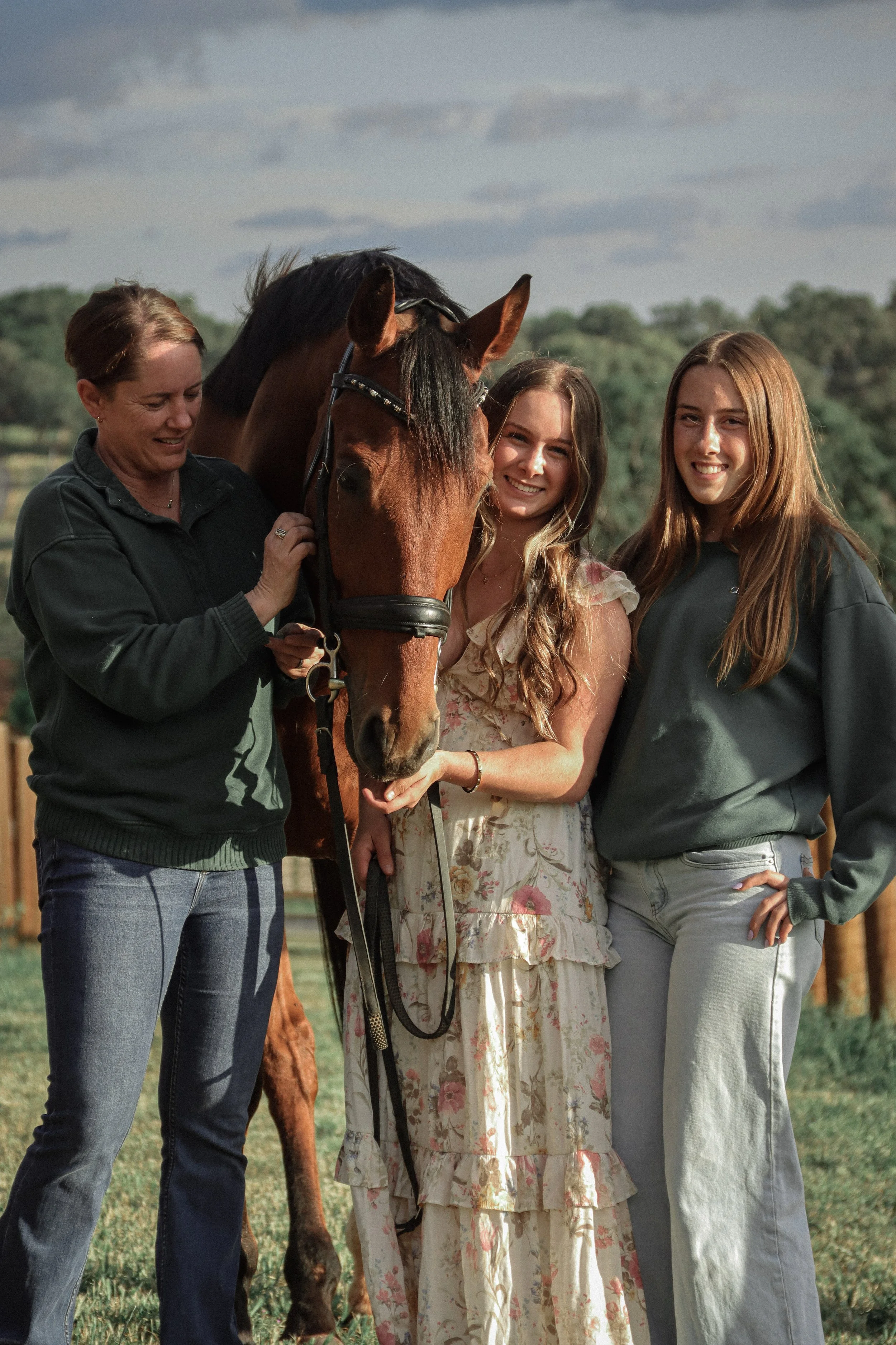 Group of four people and a horse outdoors, with trees in the background. The woman on the left is smiling and touching the horse, the two young women are standing close to the horse, one smiling and the other with a natural smile, and are dressed in 