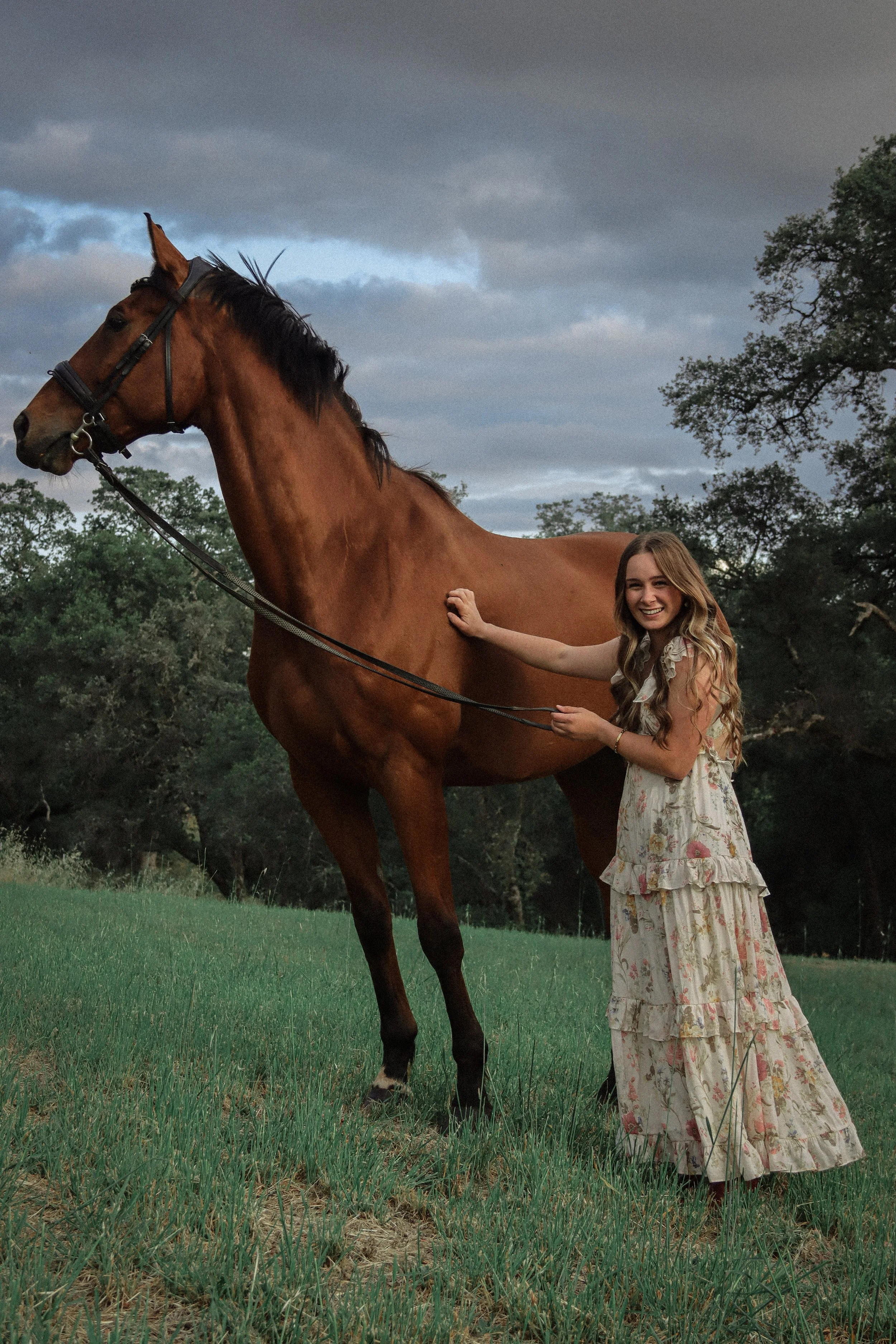 A young woman with long blonde hair wearing a floral dress petting a chestnut horse on the neck in a grassy field with trees and cloudy sky in the background.