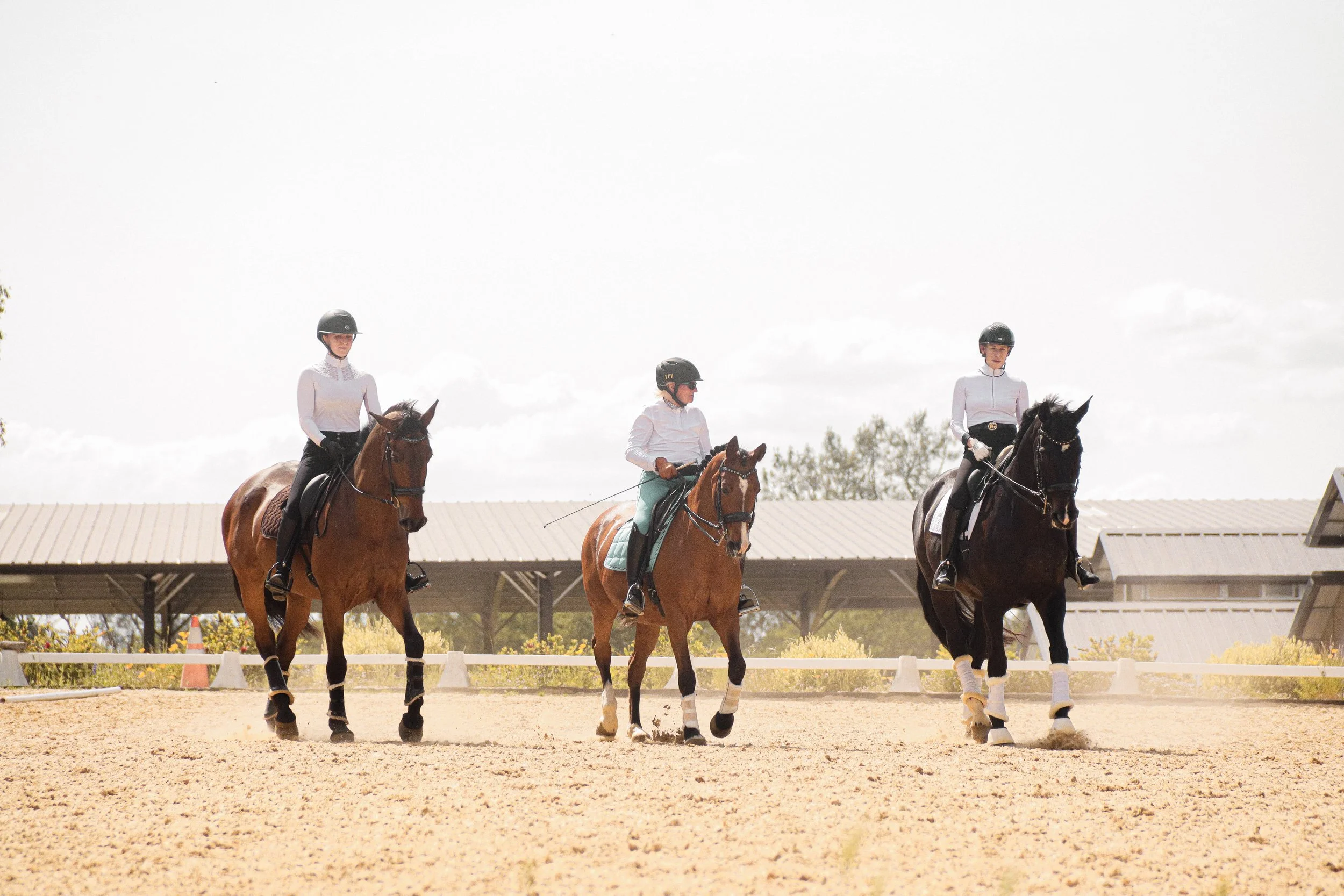 Three women in riding helmets and white shirts riding horses on a dirt arena with a roofed structure and cloudy sky in the background.