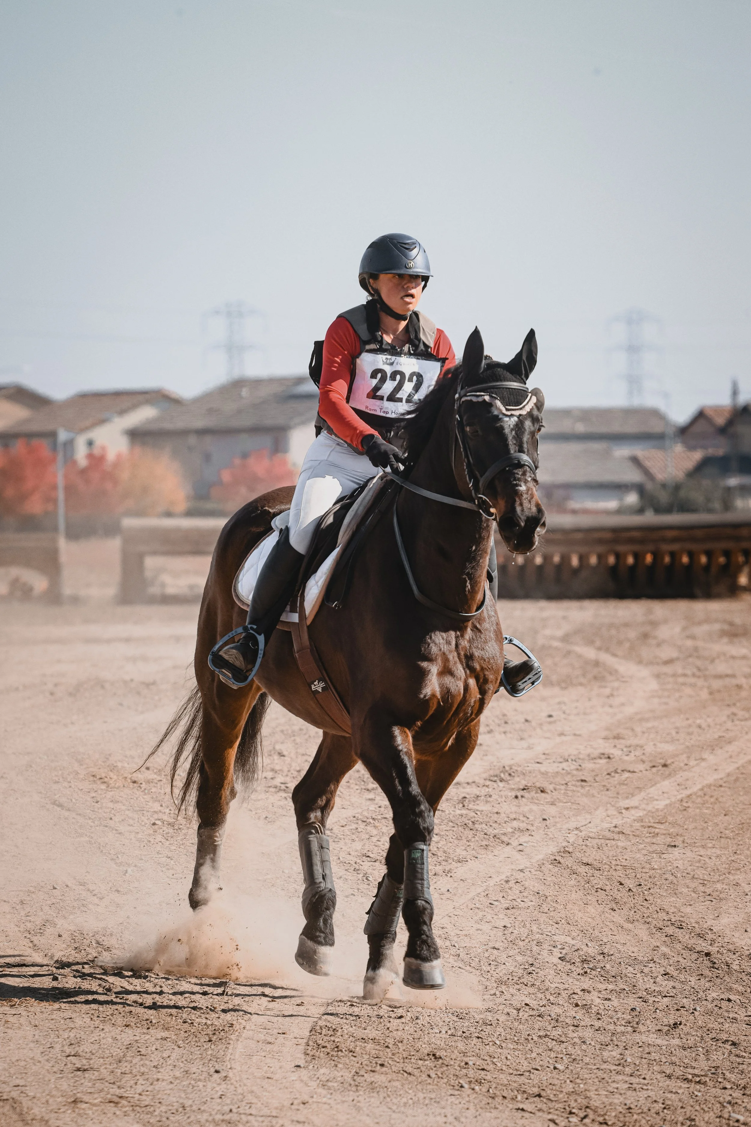 A woman wearing a helmet and riding gear, with a numbered vest, riding a brown horse on a dirt track, participating in an equestrian event.