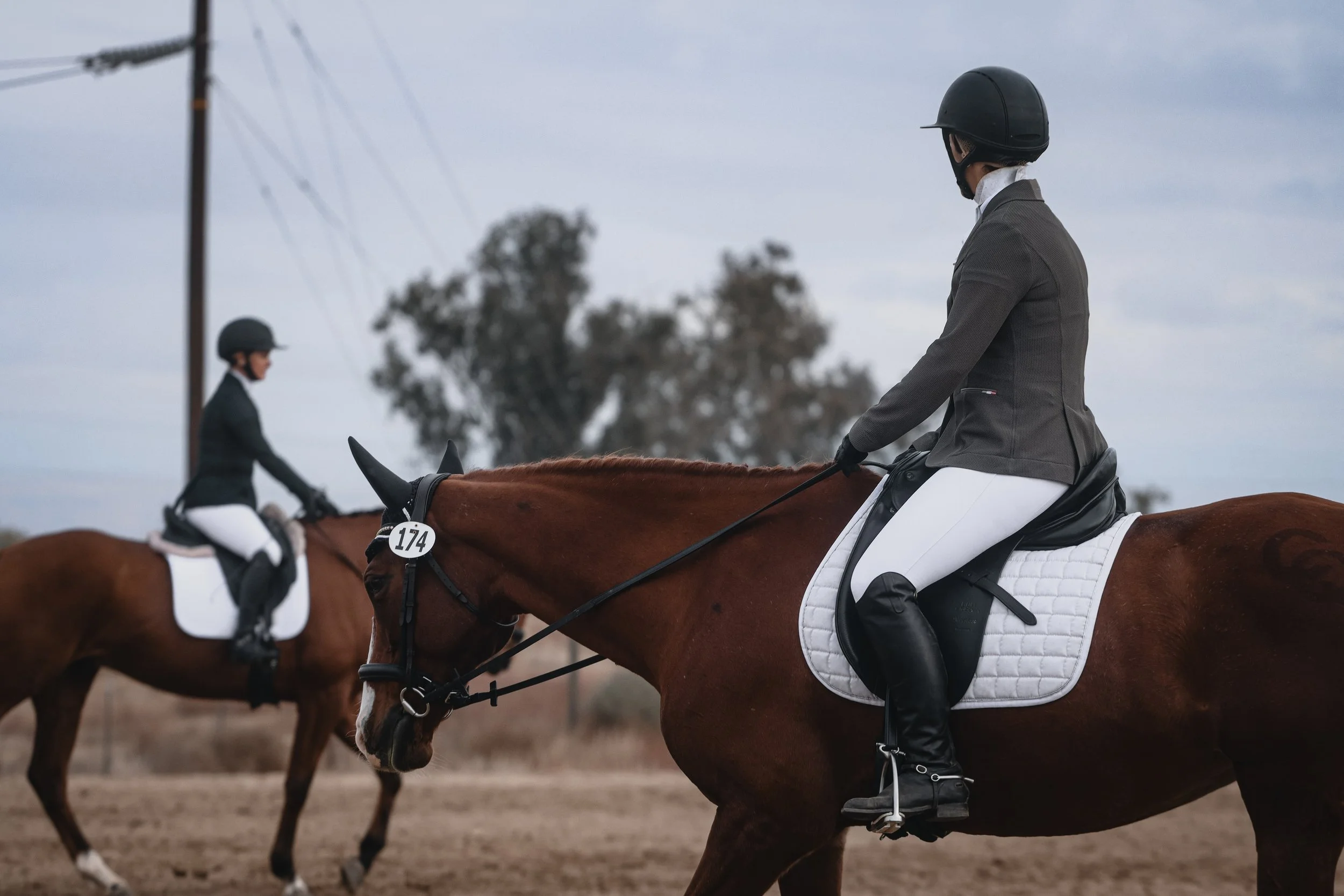 Riders in dressage attire on horseback during a competition or practice, with a focus on the rider in the foreground wearing a helmet and formal riding gear.