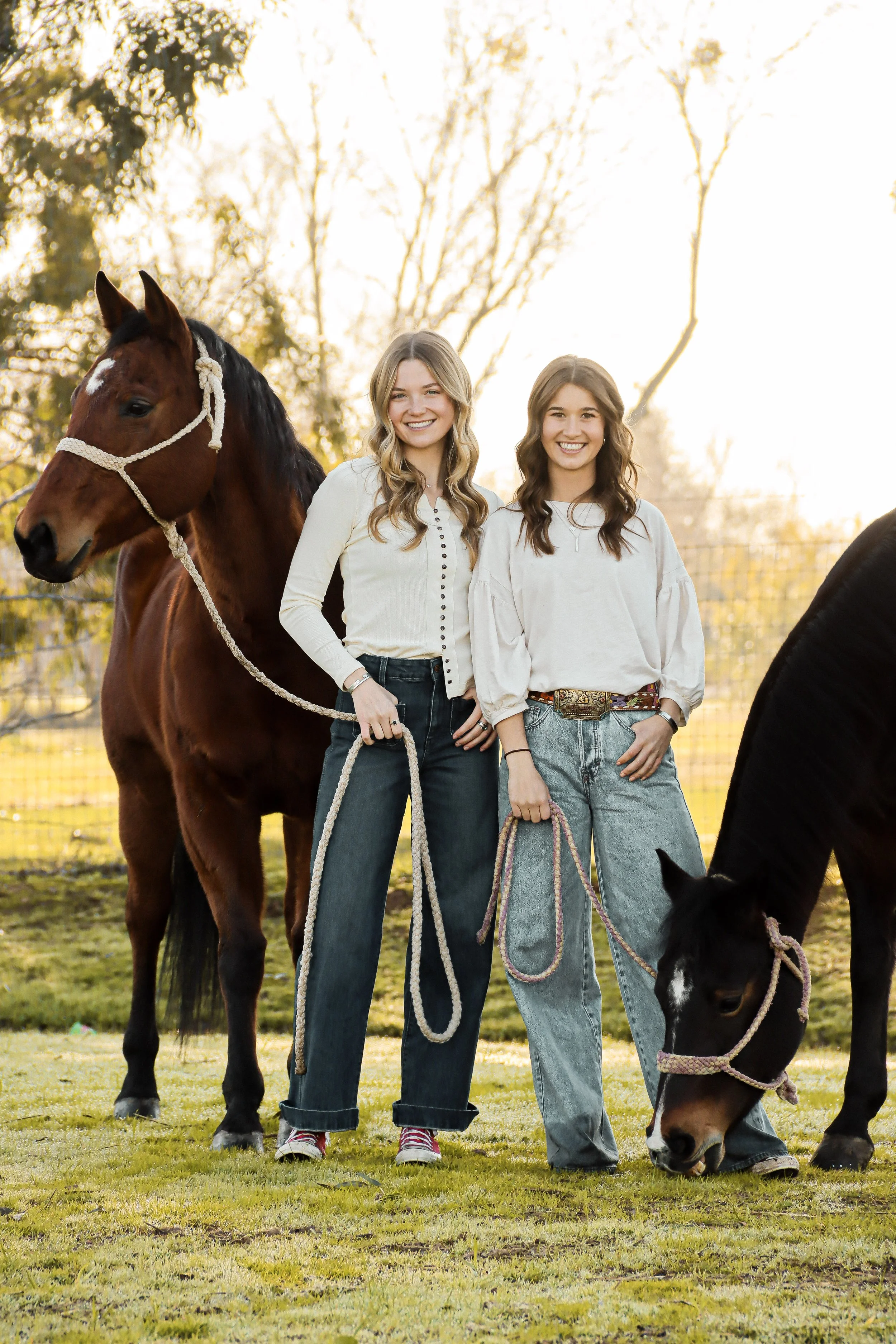 Two young women holding horse leads standing outdoors in a grassy area with trees in the background, during sunset.