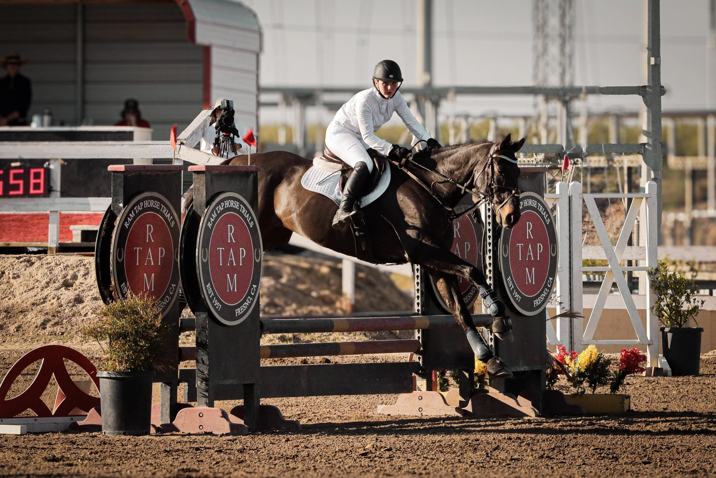 A young person wearing a riding helmet and white riding attire participating in a horse jumping competition indoors. The horse is jumping over a colorful obstacle with the words "R T M TAP" on it, and a camera operator is capturing the jump.