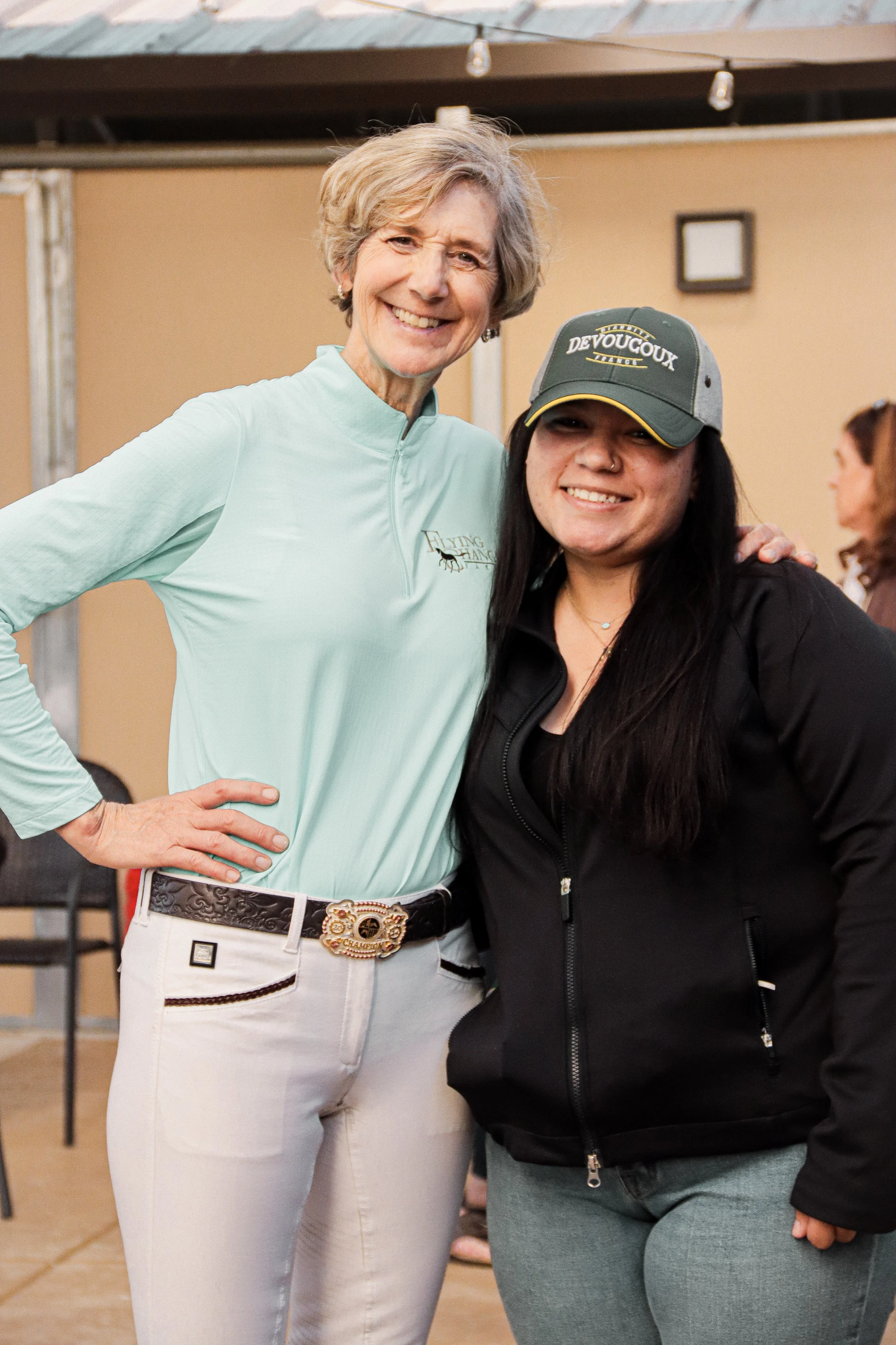 Two women smiling, standing close together indoors. One woman has short gray hair, is wearing a light green long-sleeve shirt with a logo and white pants with a belt. The other woman has long dark hair, is wearing a black jacket with a zipper, a cap 