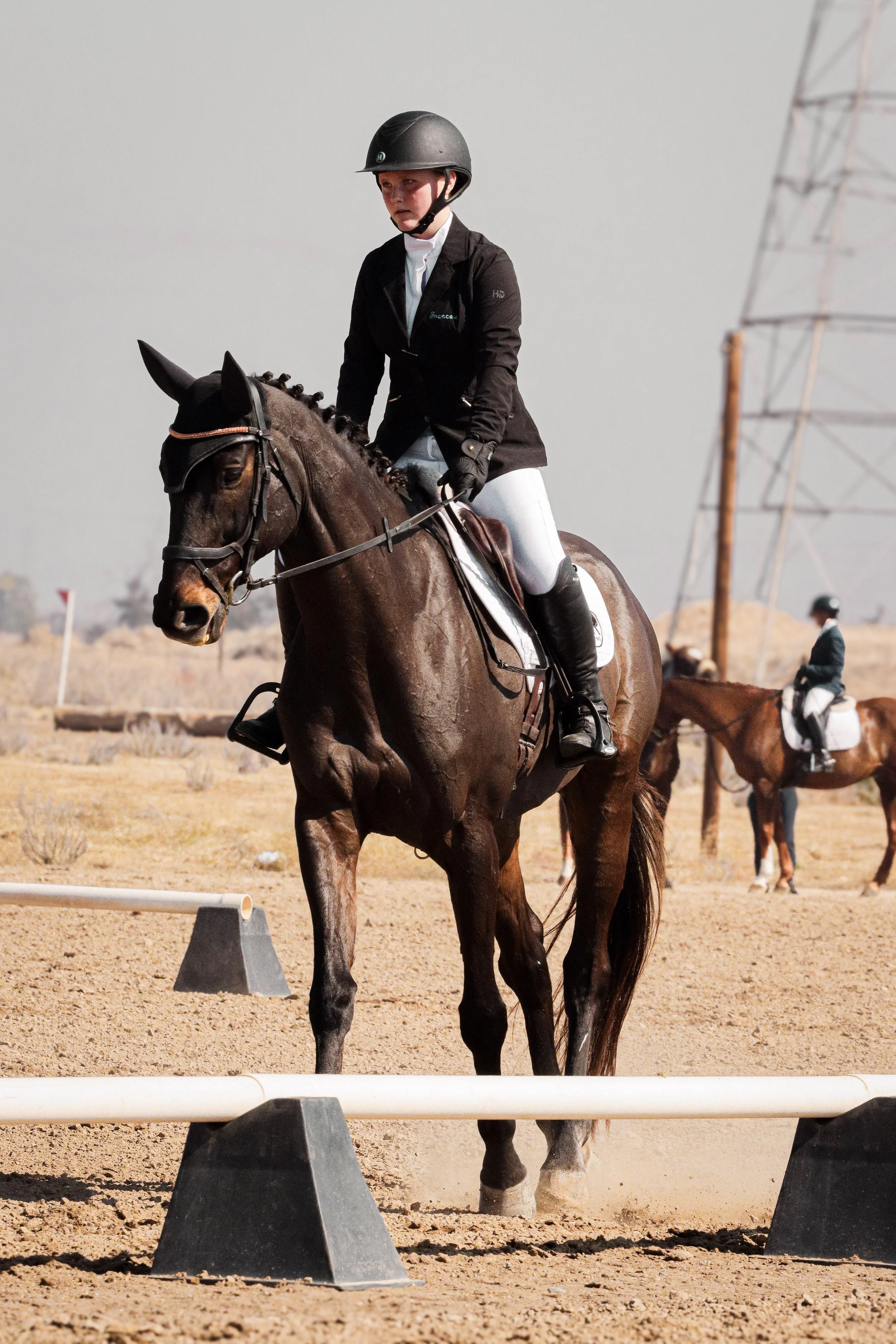 Young boy riding a horse in an outdoor equestrian arena, wearing a black helmet, black jacket, and white riding pants.