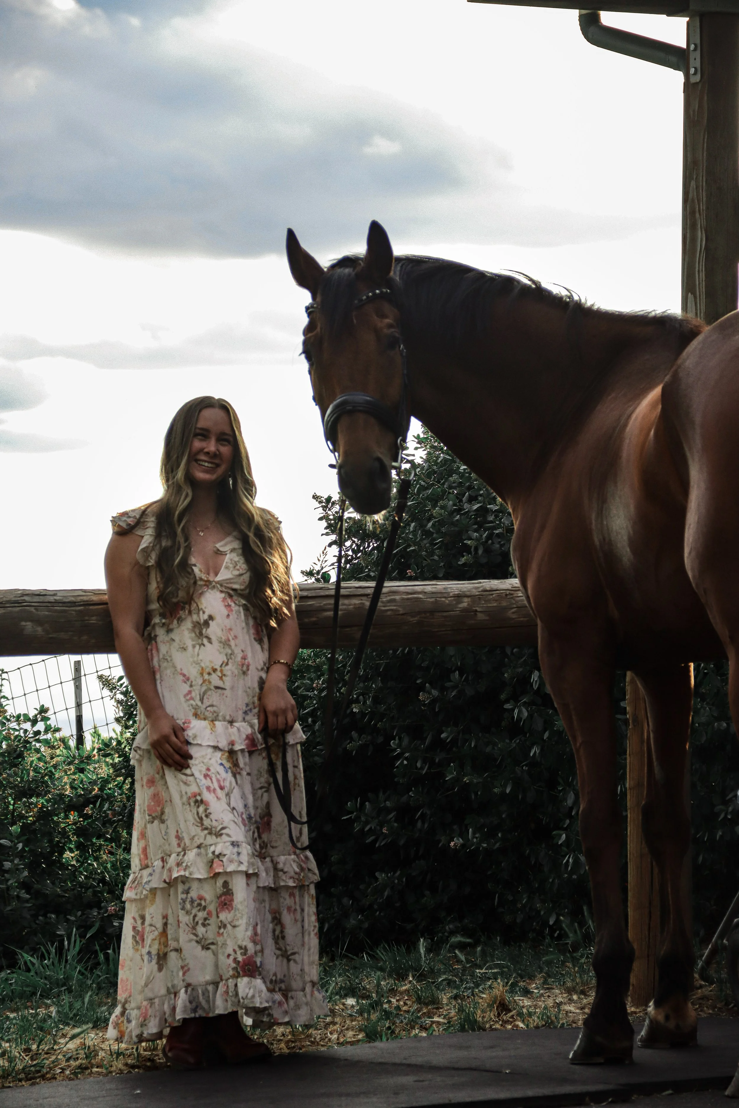 A young woman with long wavy hair wearing a floral dress, standing outdoors next to a brown horse with a black mane, under a cloudy sky.