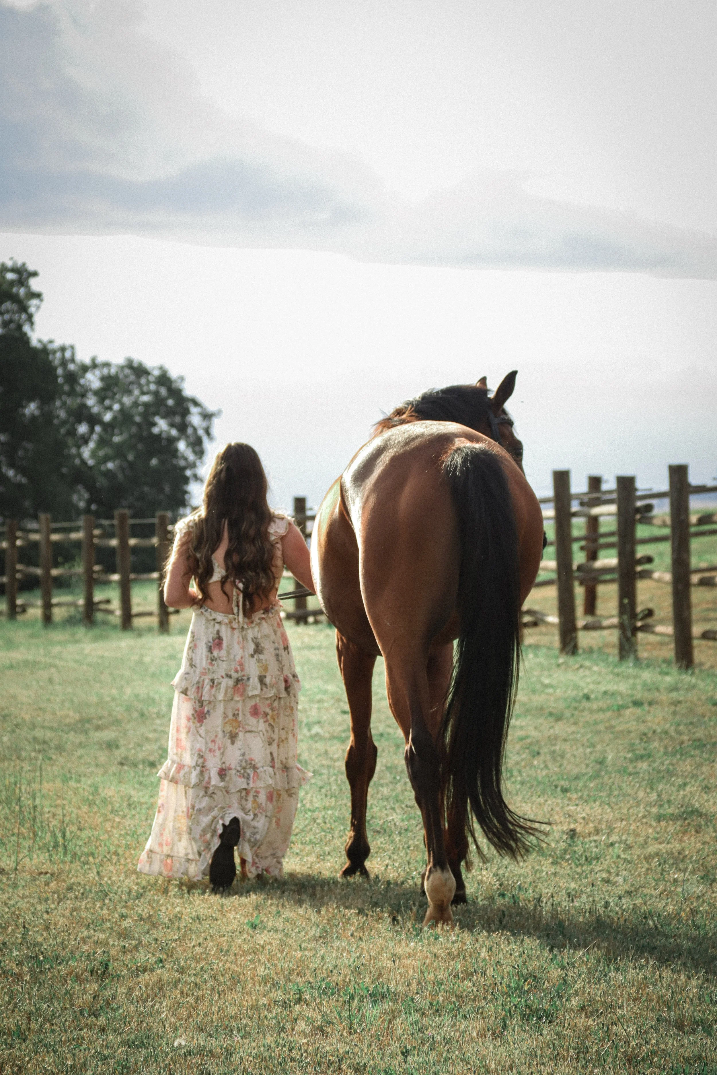A woman with long, wavy brown hair in a floral dress walks beside a brown horse with a black mane and tail in a fenced grassy field under a cloudy sky.